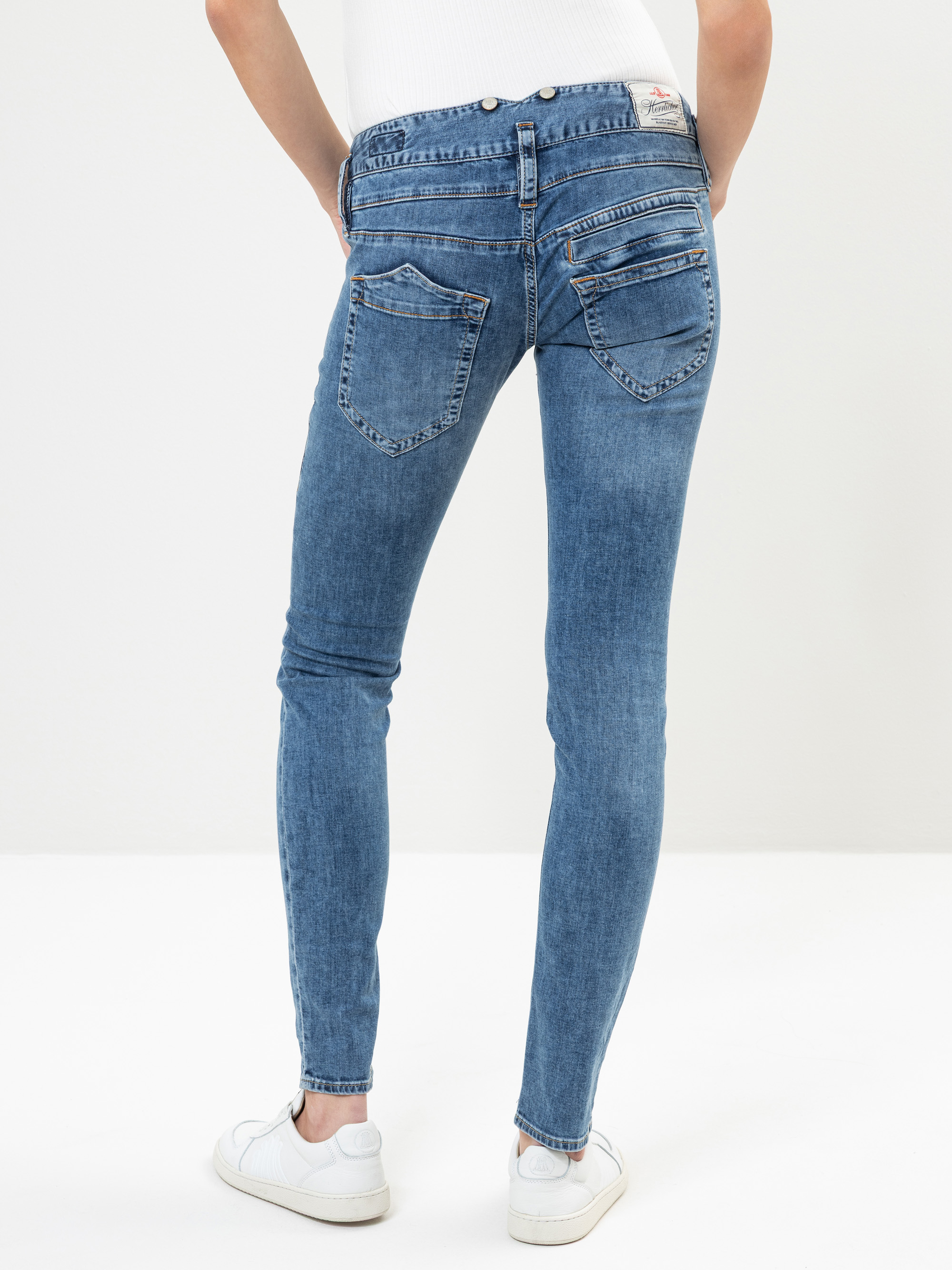 Eine Person in hellblauen Skinny-Jeans und weißen Turnschuhen steht mit den Händen in den Hosentaschen vor einem schlichten weißen Hintergrund.