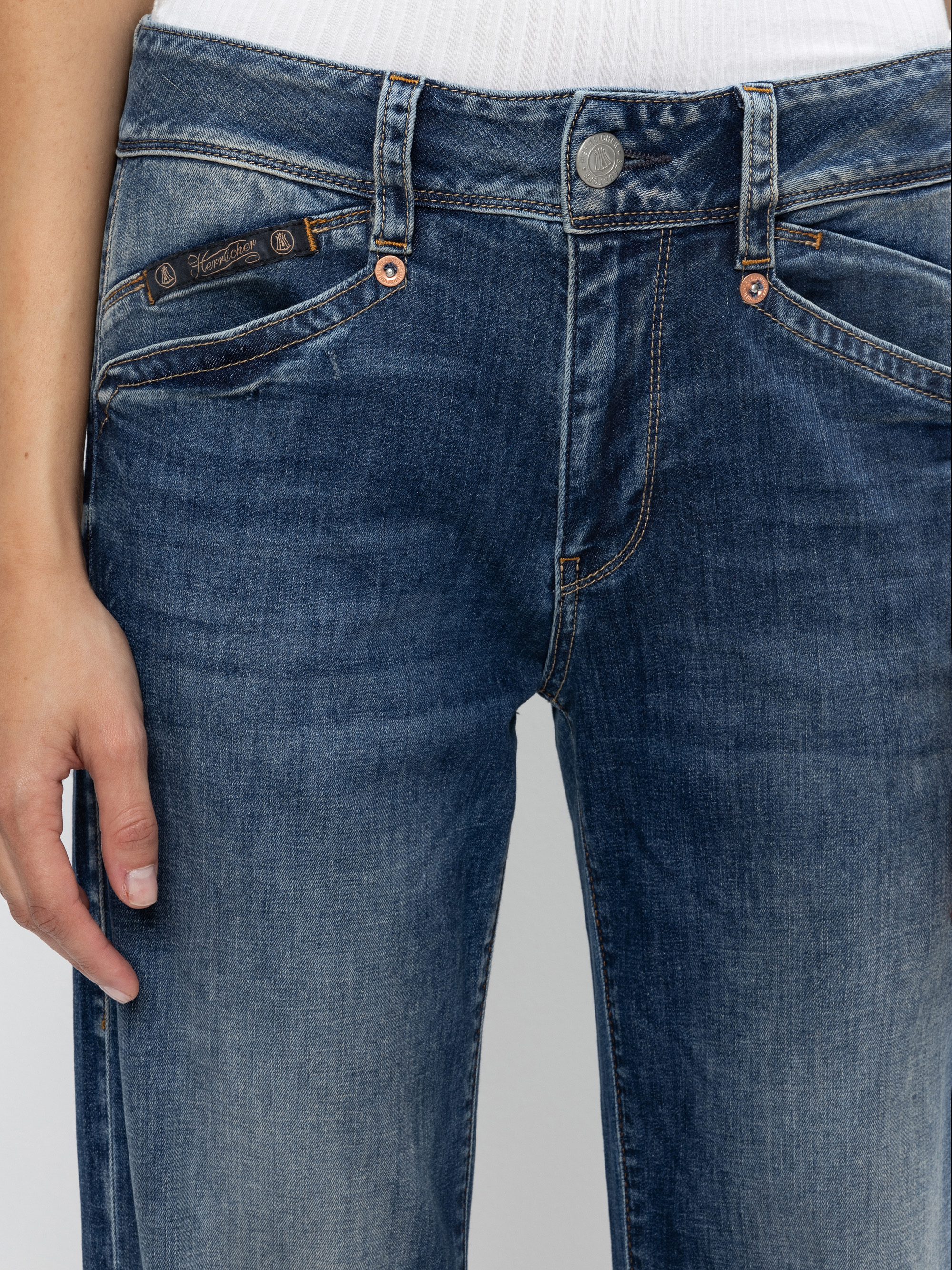 Eine Person, die blaue Jeans und ein weißes Oberteil trägt, von der Taille bis zur Mitte des Oberschenkels, mit einer sichtbaren Hand an der Seite liegend.