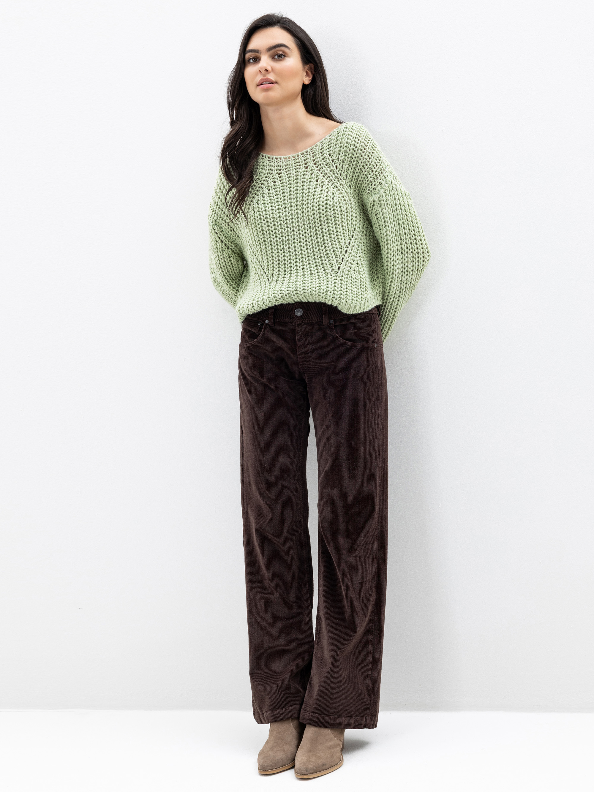 Eine Frau steht vor einer weißen Wand und trägt einen lockeren, hellgrünen Strickpullover, eine hoch sitzende braune Cordhose mit weitem Bein und beigefarbene Stiefeletten. Sie hat langes dunkles Haar und einen entspannten Gesichtsausdruck.