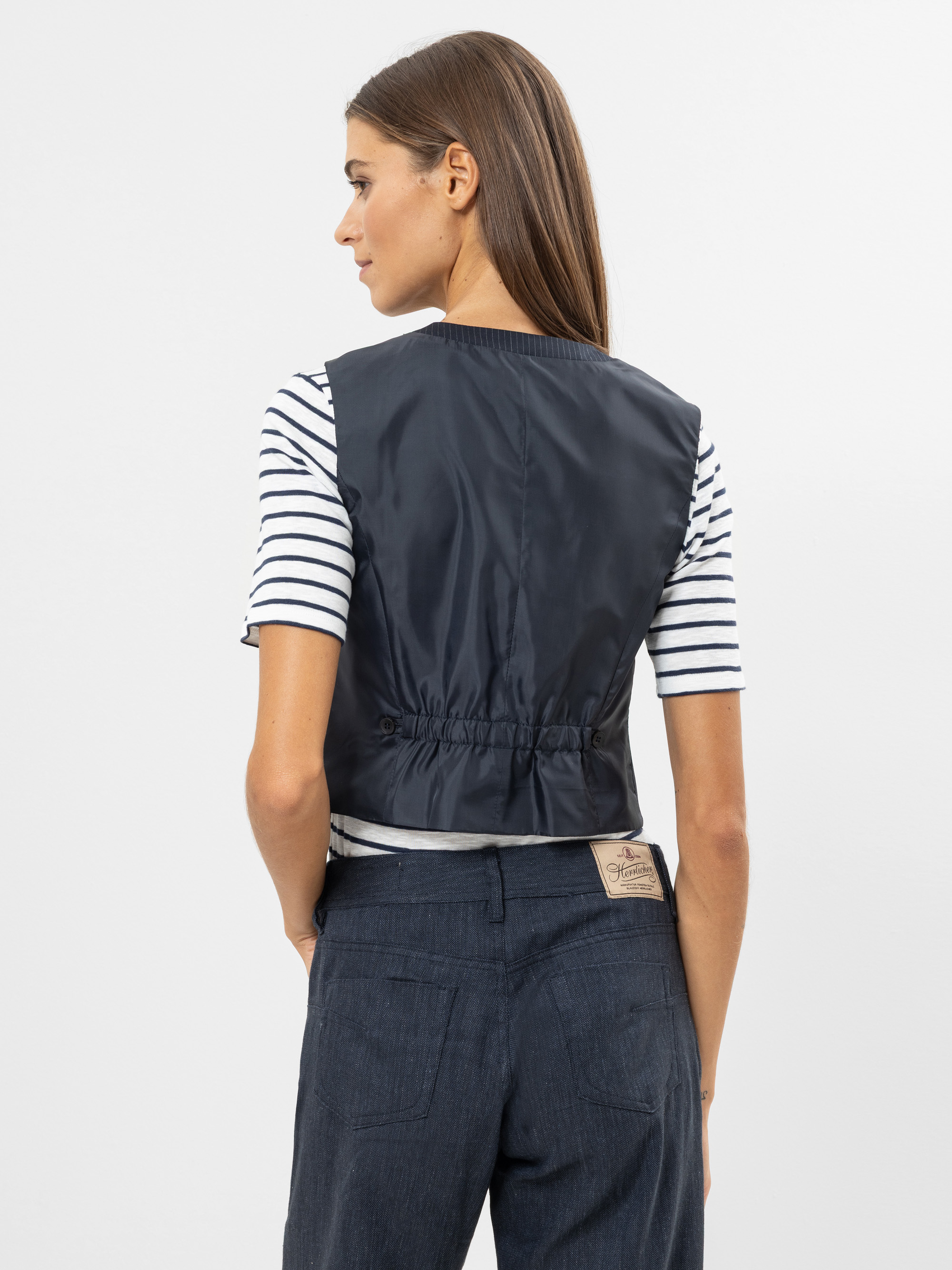 Eine Frau mit glattem braunem Haar, die eine schwarze ärmellose Weste über einem weiß-marinefarbenen gestreiften Hemd und dunkelblaue Jeans trägt, steht mit dem Gesicht zur Kamera vor einem schlichten hellen Hintergrund.