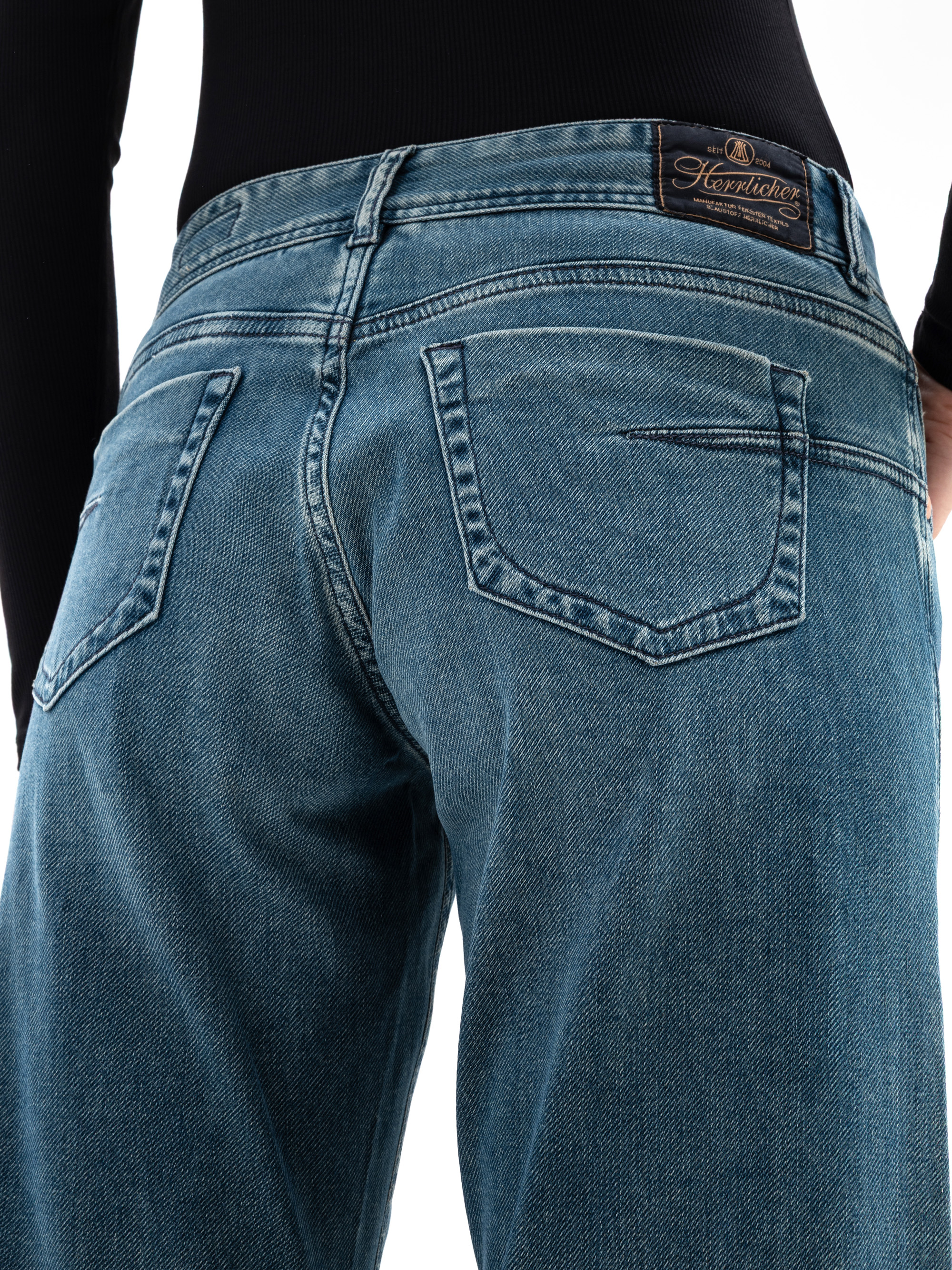 Nahaufnahme einer Person, die eine blaue Denim-Jeans und ein schwarzes langärmeliges Oberteil trägt, wobei die Gesäßtaschen und ein Leder-Markenaufnäher am Bund zu sehen sind.