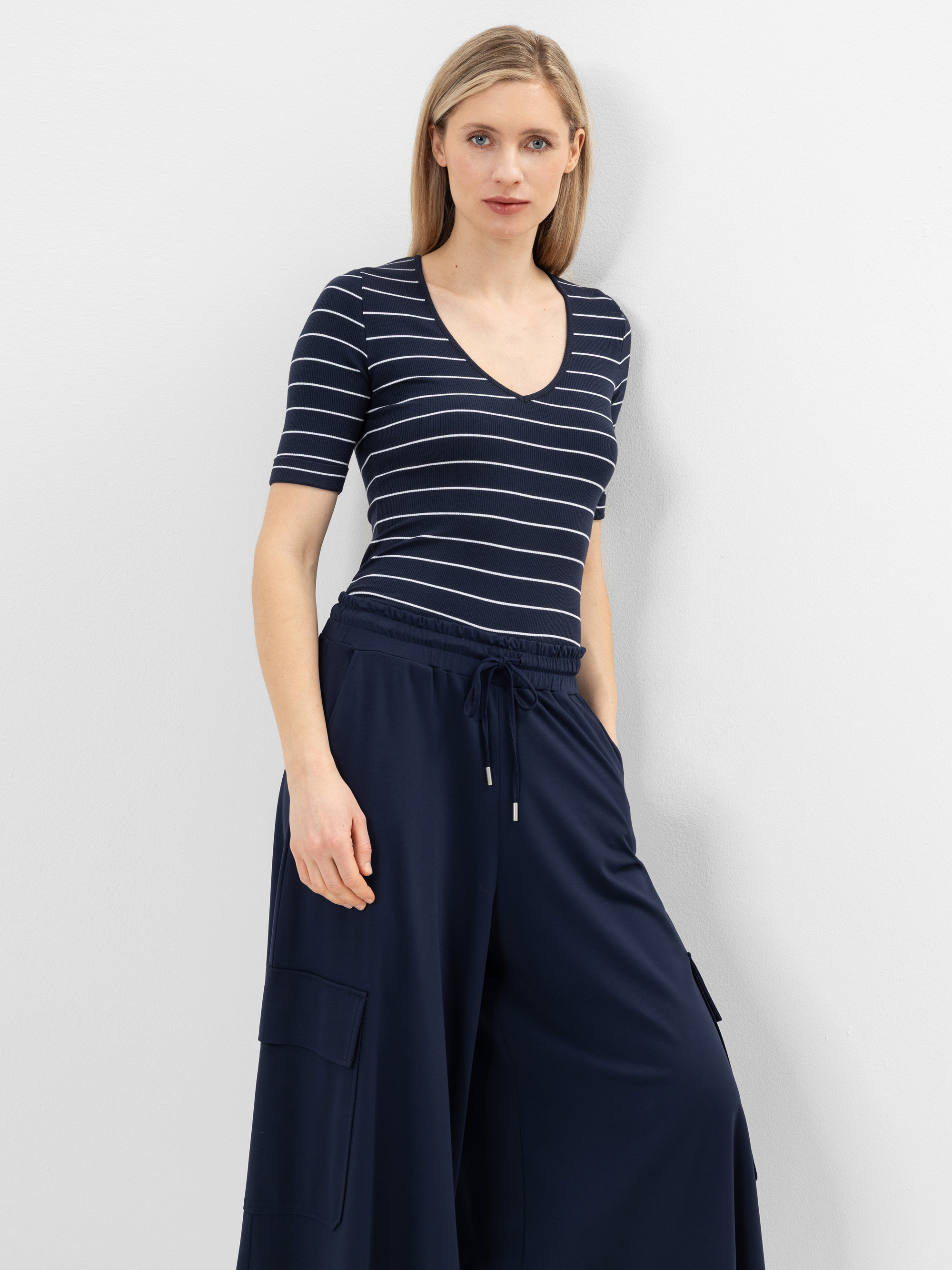 Eine Frau mit glattem blondem Haar trägt ein marineblaues, kurzärmeliges Oberteil mit V-Ausschnitt und weißen Querstreifen sowie eine passende marineblaue Hose mit weiten Beinen und Taschen. Sie steht vor einer schlichten weißen Wand und schaut in die