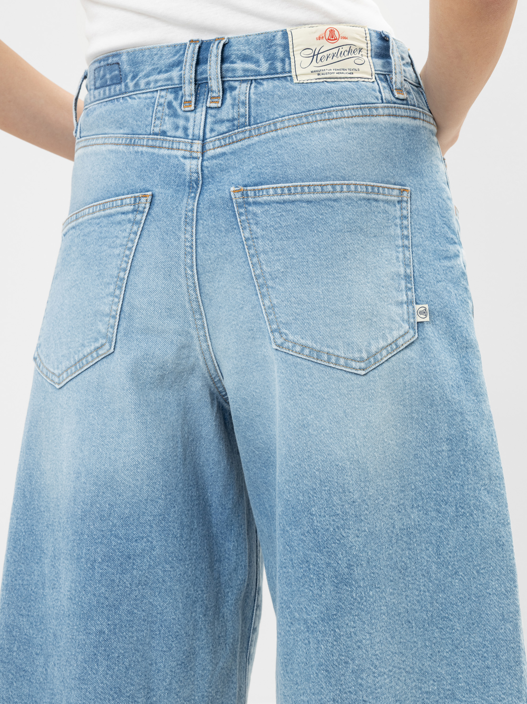 Eine Person trägt eine hellblaue Jeans mit weitem Bein, von hinten gesehen, mit zwei großen Gesäßtaschen und einem weißen Heritage-Aufnäher am Bund.