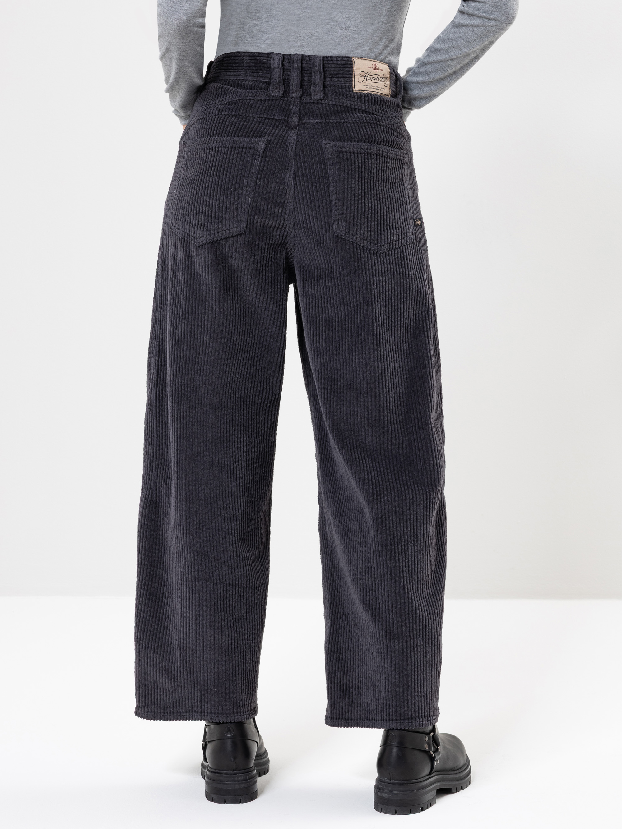 Eine Person steht mit dem Gesicht nach hinten, trägt eine dunkelgraue, weit geschnittene Cordhose, ein graues, langärmeliges Oberteil und schwarze Stiefel vor einem schlichten weißen Hintergrund.
