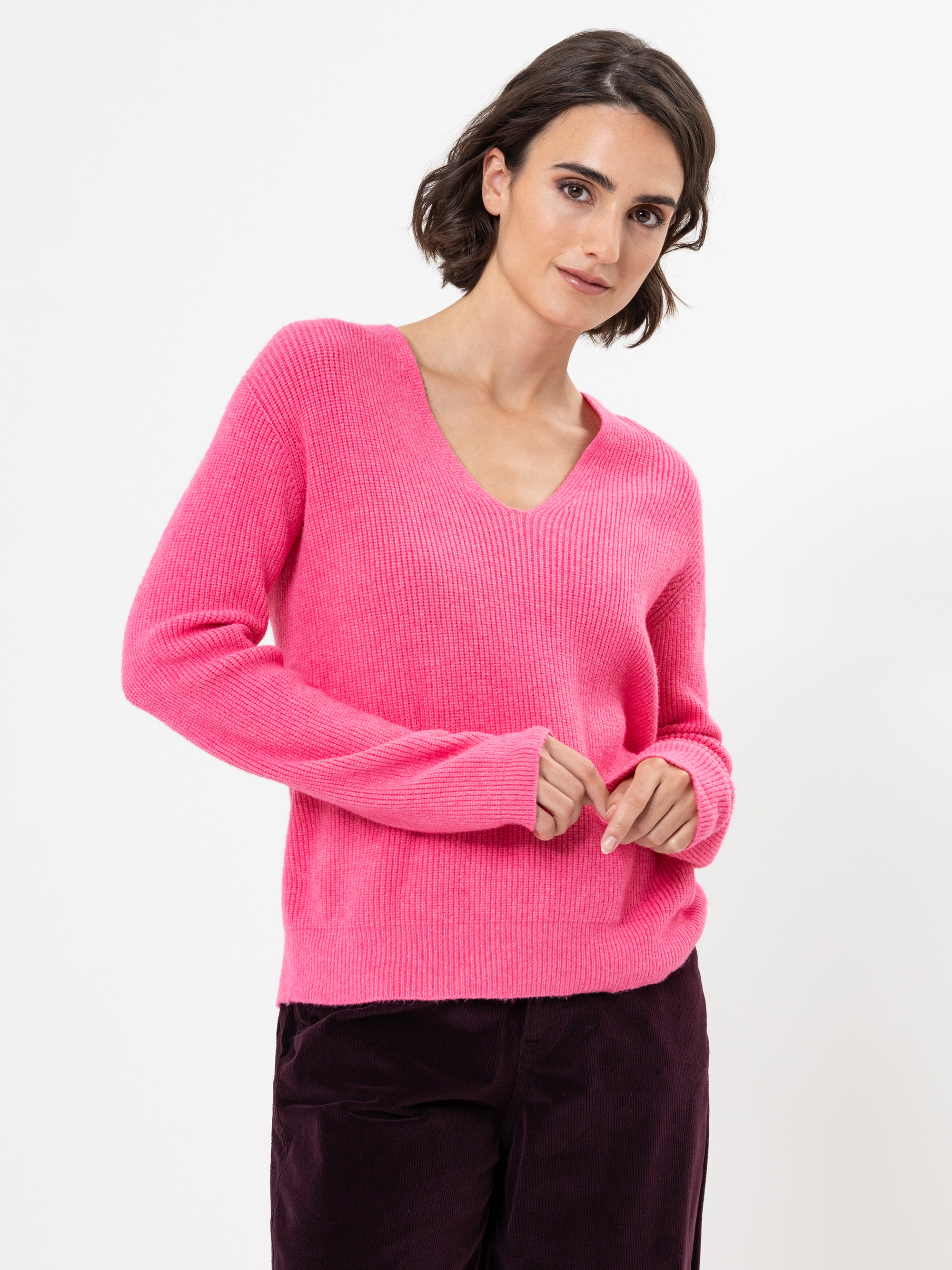 Frau mit kurzen braunen Haaren, die einen hellrosa Pullover mit V-Ausschnitt und eine dunkelviolette Hose trägt und vor einem schlichten weißen Hintergrund steht.