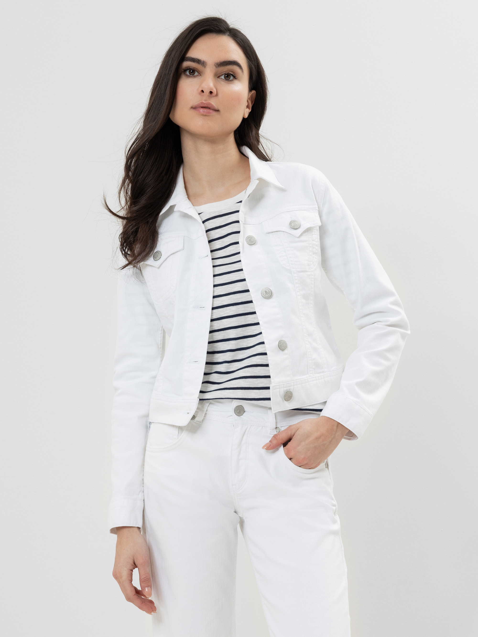 Eine Frau mit langen dunklen Haaren trägt eine weiße Jeansjacke über einem weiß-marinefarbenen gestreiften Hemd und einer weißen Hose. Sie steht vor einem schlichten hellen Hintergrund und hat einen neutralen Gesichtsausdruck.