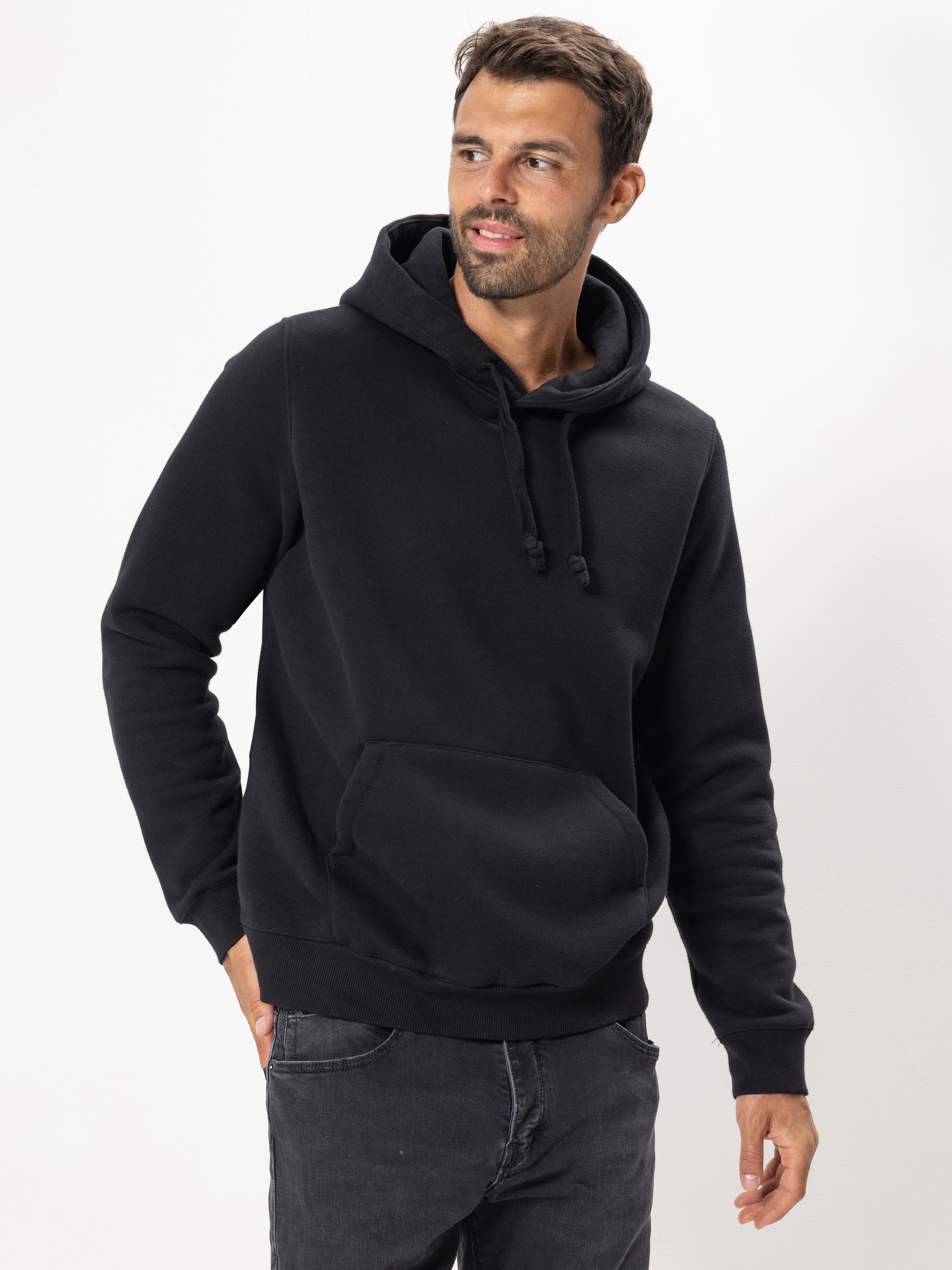 Ein Mann mit schwarzem Kapuzenpulli und dunkler Jeans steht vor einem schlichten weißen Hintergrund und blickt mit entspannter Miene leicht nach links.