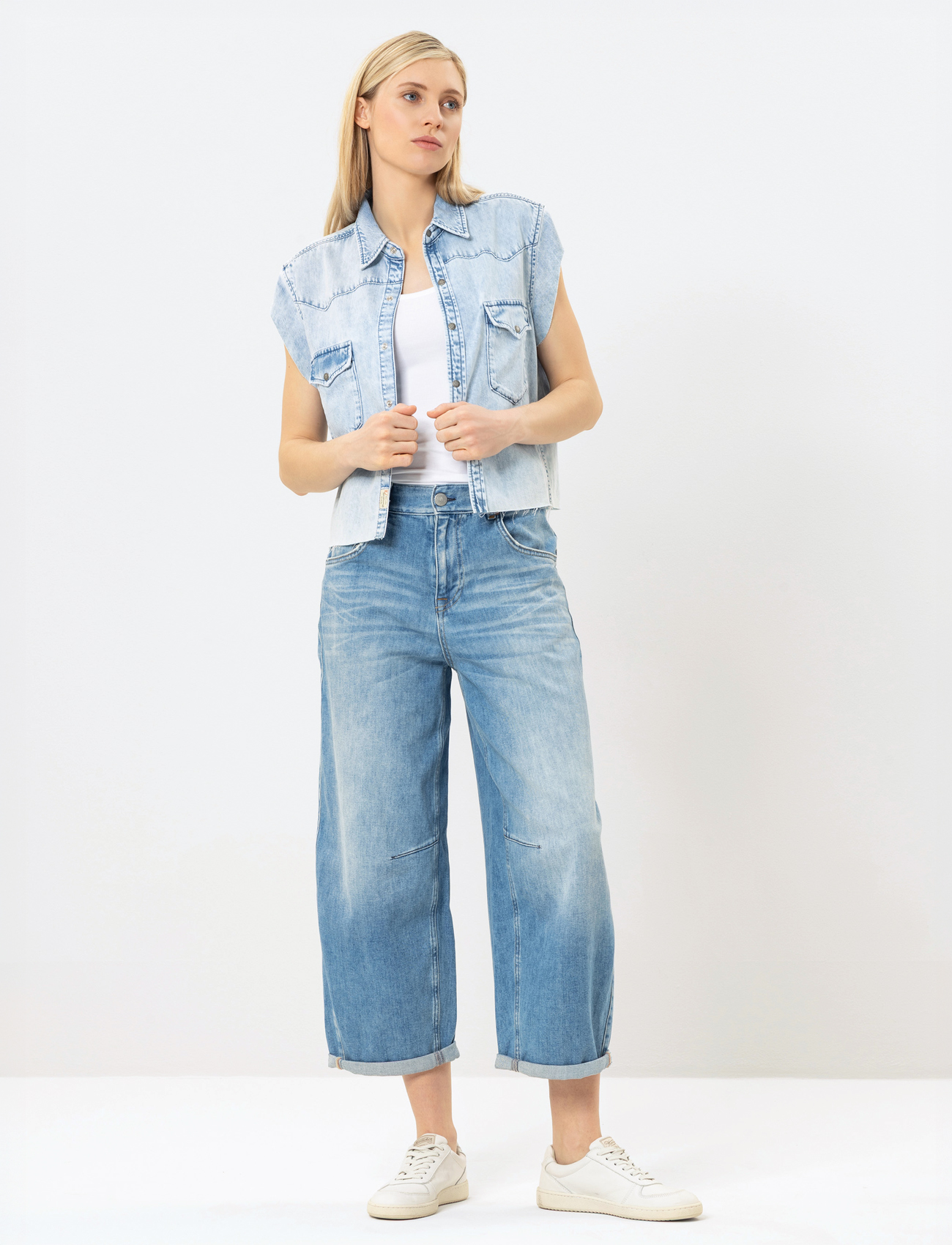Eine Frau steht vor einem schlichten Hintergrund und trägt ein ärmelloses Jeanshemd über einem weißen Oberteil, hellblaue Jeans mit weitem Bein und weiße Turnschuhe. Sie hat langes blondes Haar und einen neutralen Gesichtsausdruck.