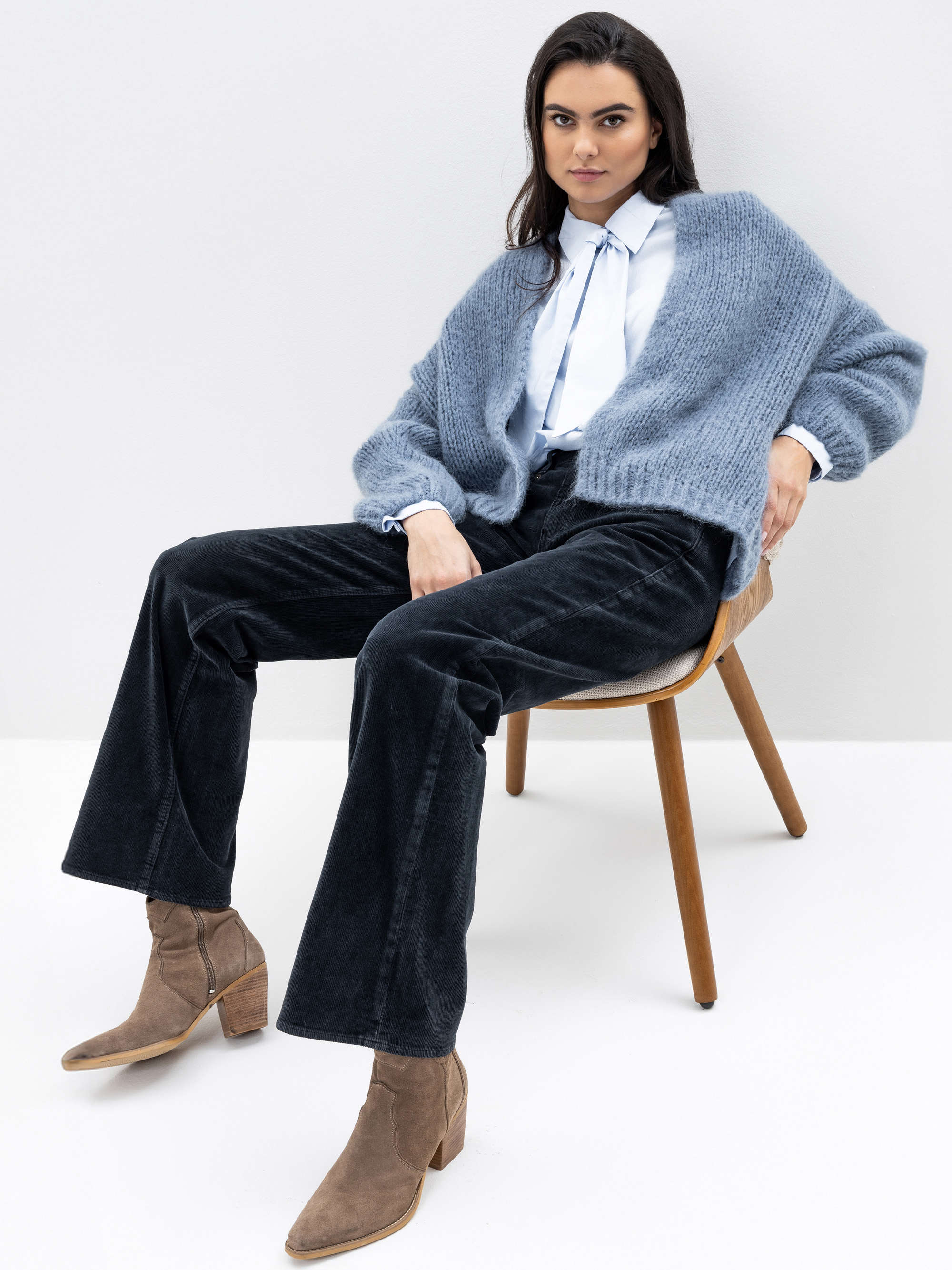 Eine Frau sitzt auf einem Holzstuhl, trägt eine hellblaue Strickjacke über einem weißen Hemd, eine schwarze Hose mit weitem Bein und braunen Stiefeln mit Absätzen und blickt selbstbewusst in die Kamera vor einem schlichten Hintergrund.