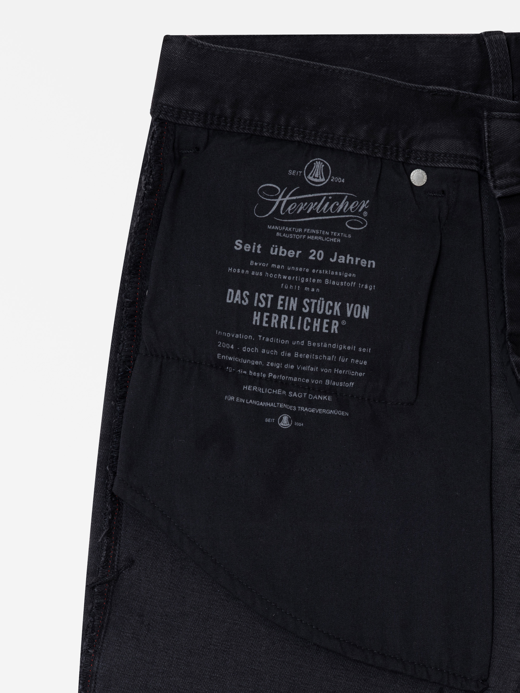 Nahaufnahme der Innentasche einer schwarzen Hose mit einem gedruckten Etikett in deutscher Sprache, auf dem die Marke Herrlicher, ihre über 20-jährige Geschichte und ein Dankeschön für die Wahl ihres Qualitätsprodukts angegeben sind.