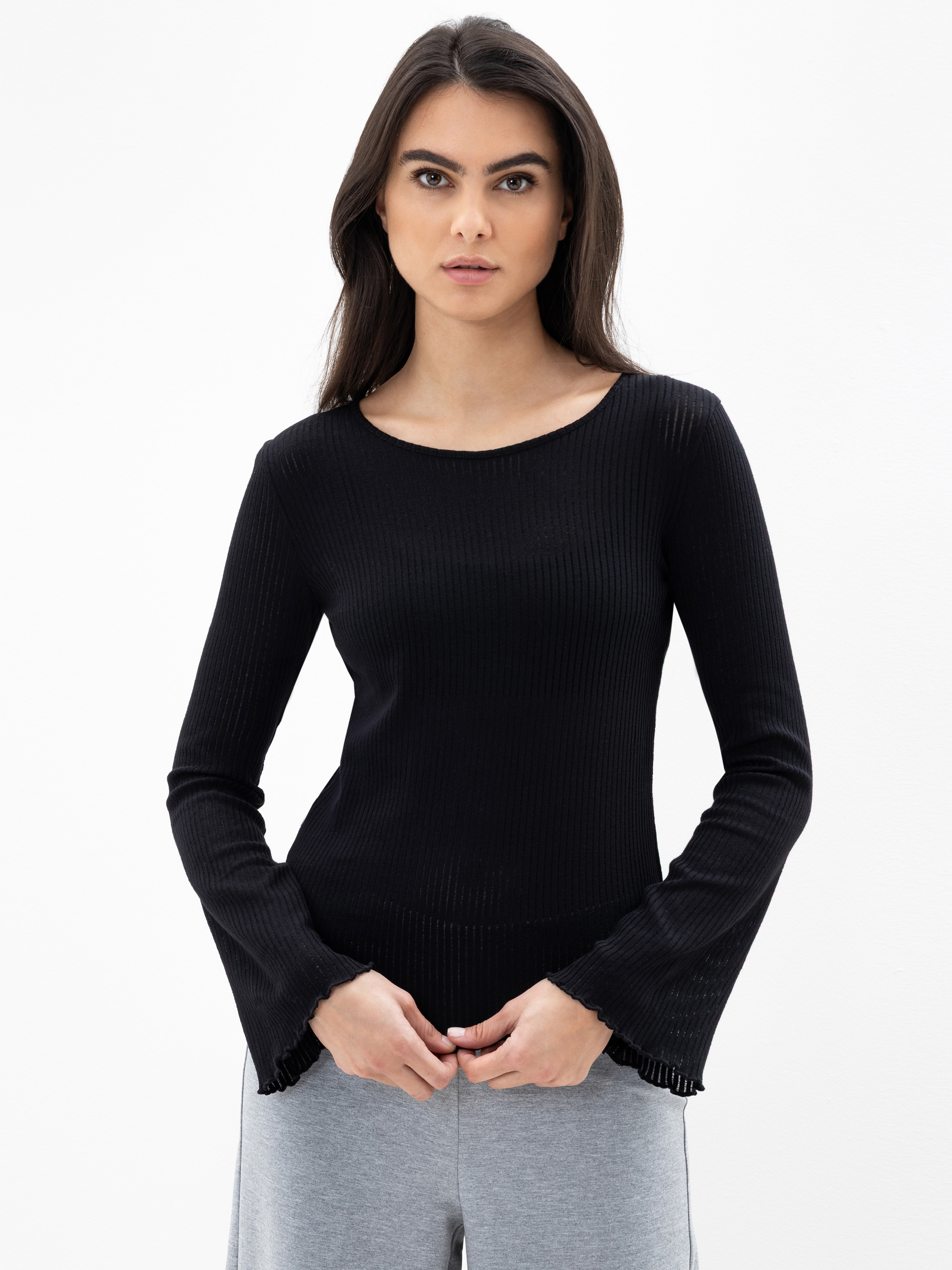 Eine Frau mit langen dunklen Haaren trägt ein schwarzes, langärmeliges, geripptes Strickoberteil und eine hellgraue Hose. Sie steht vor einem schlichten weißen Hintergrund und blickt mit neutralem Gesichtsausdruck direkt in die Kamera.