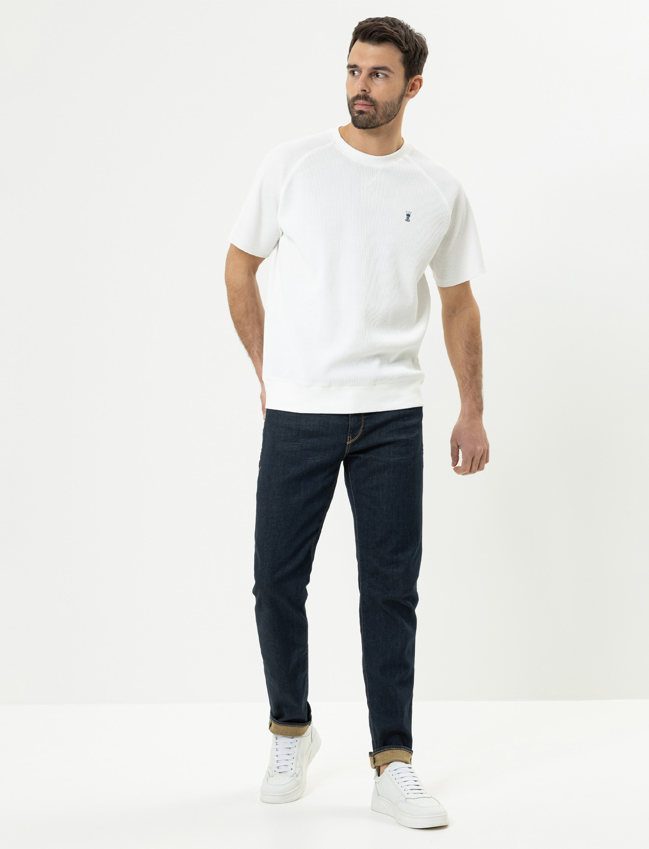 Ein Mann mit Bart, der ein weißes kurzärmeliges Hemd, dunkelblaue Jeans und weiße Turnschuhe trägt, steht vor einem schlichten weißen Hintergrund. Er blickt nach links und hat eine Hand in der Tasche.