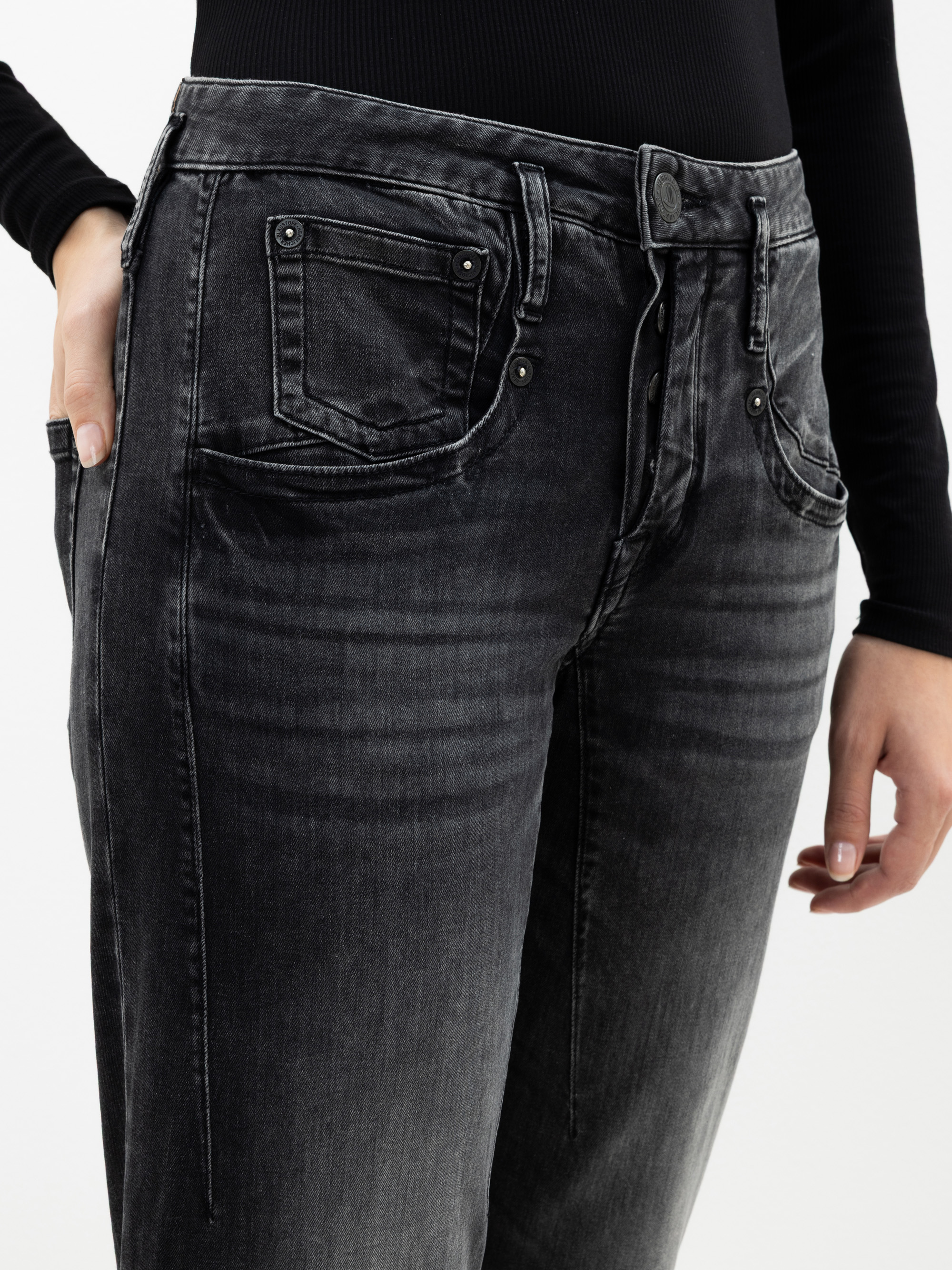 Eine Person, die eine schwarze verwaschene Jeans und ein schwarzes langärmeliges Oberteil trägt, eine Hand in der Hosentasche, die andere an der Seite liegend. Das Foto zeigt den unteren Teil des Oberkörpers und die Oberschenkel.