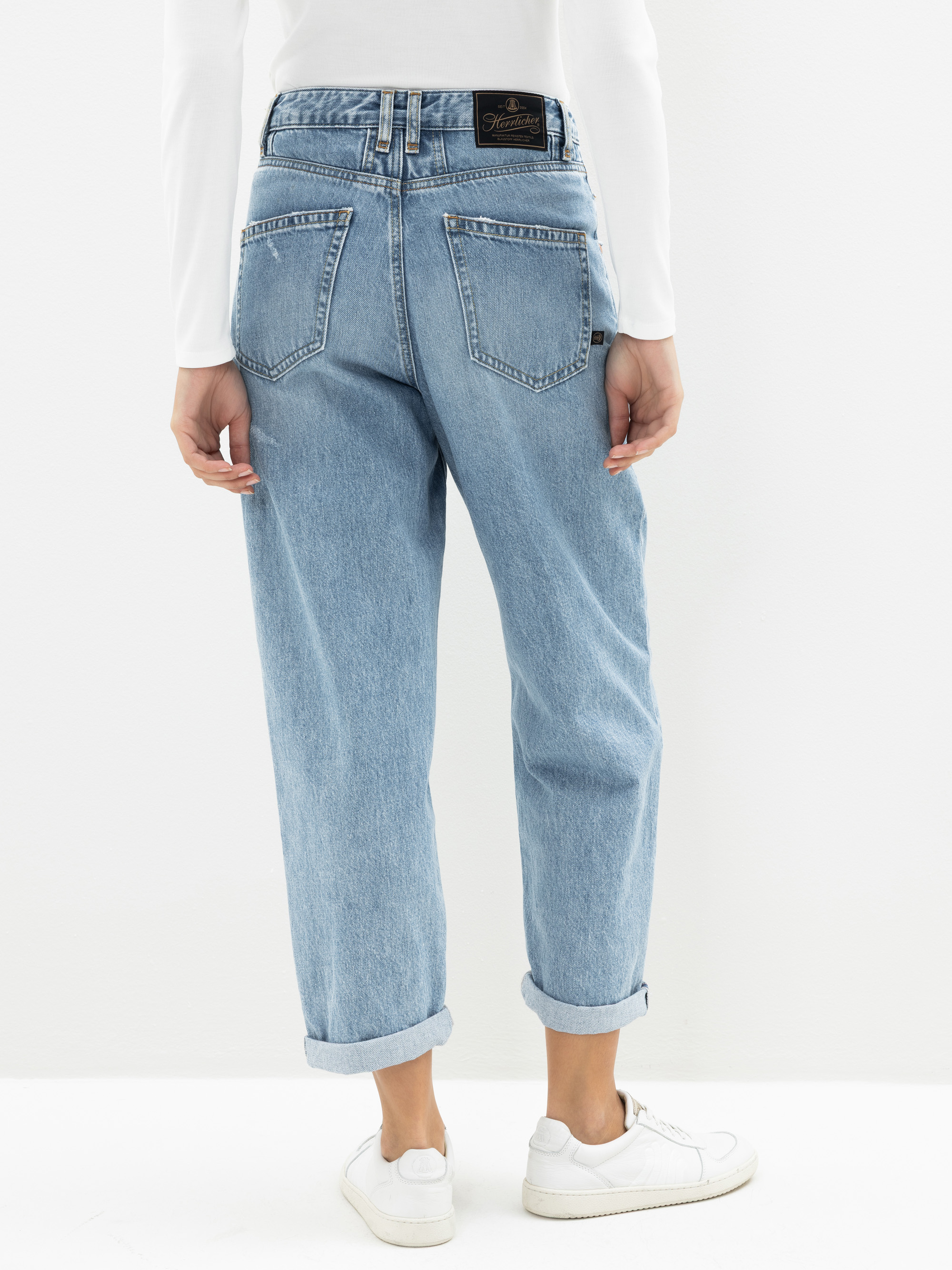 Person, die hellblaue, hoch taillierte, locker sitzende Jeans mit aufgerollten Manschetten, weiße Turnschuhe und ein weißes langärmeliges Oberteil trägt, stehend vor einem schlichten weißen Hintergrund, von hinten gesehen.