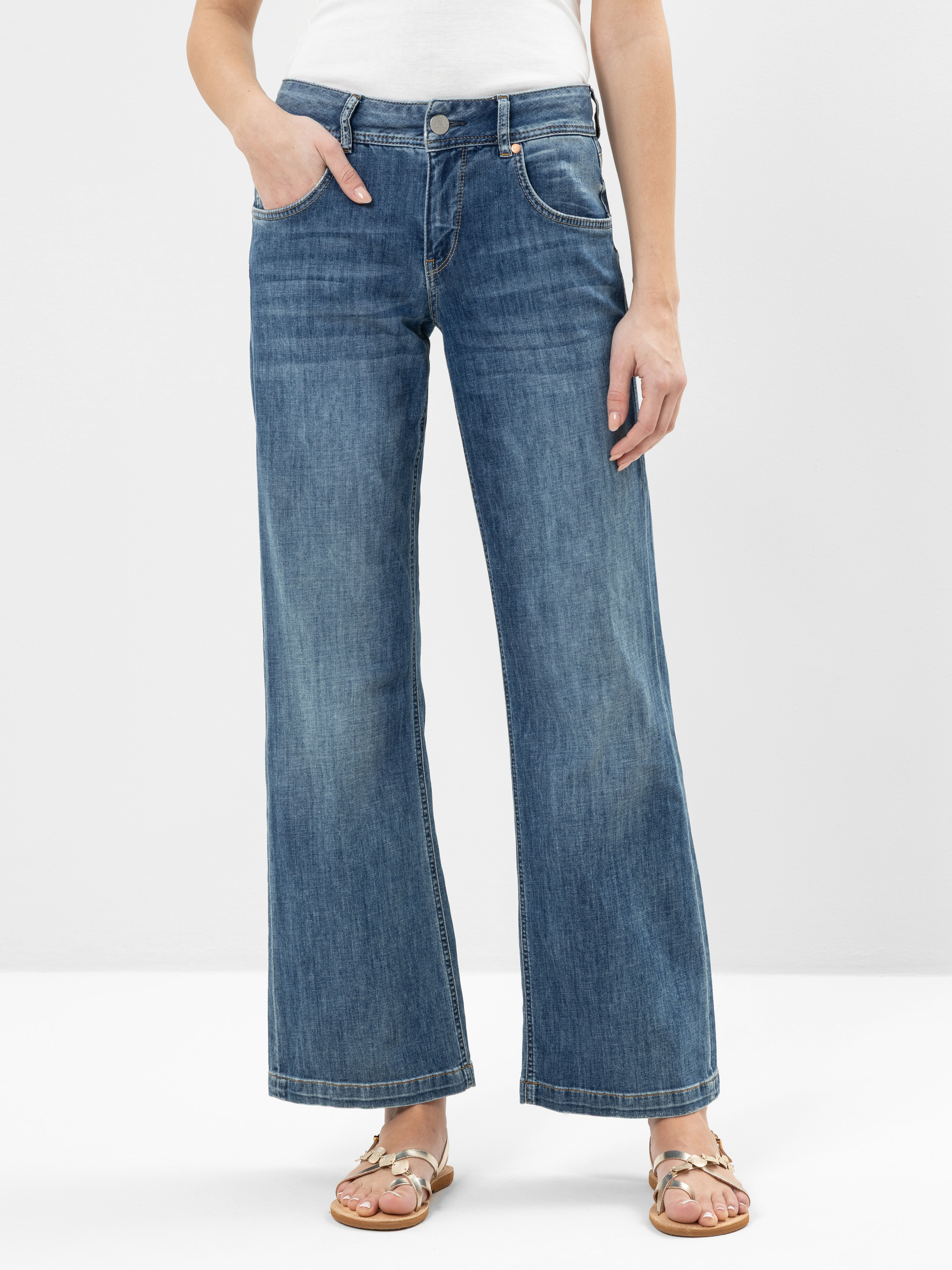 Eine Person steht vor einem schlichten Hintergrund und trägt eine blaue Jeans mit weitem Bein, ein weißes Oberteil und hellbraune Sandalen mit gekreuzten Riemen. Die Hände der Person sind entspannt, eine steckt in einer Tasche. Das Gesicht ist nicht si