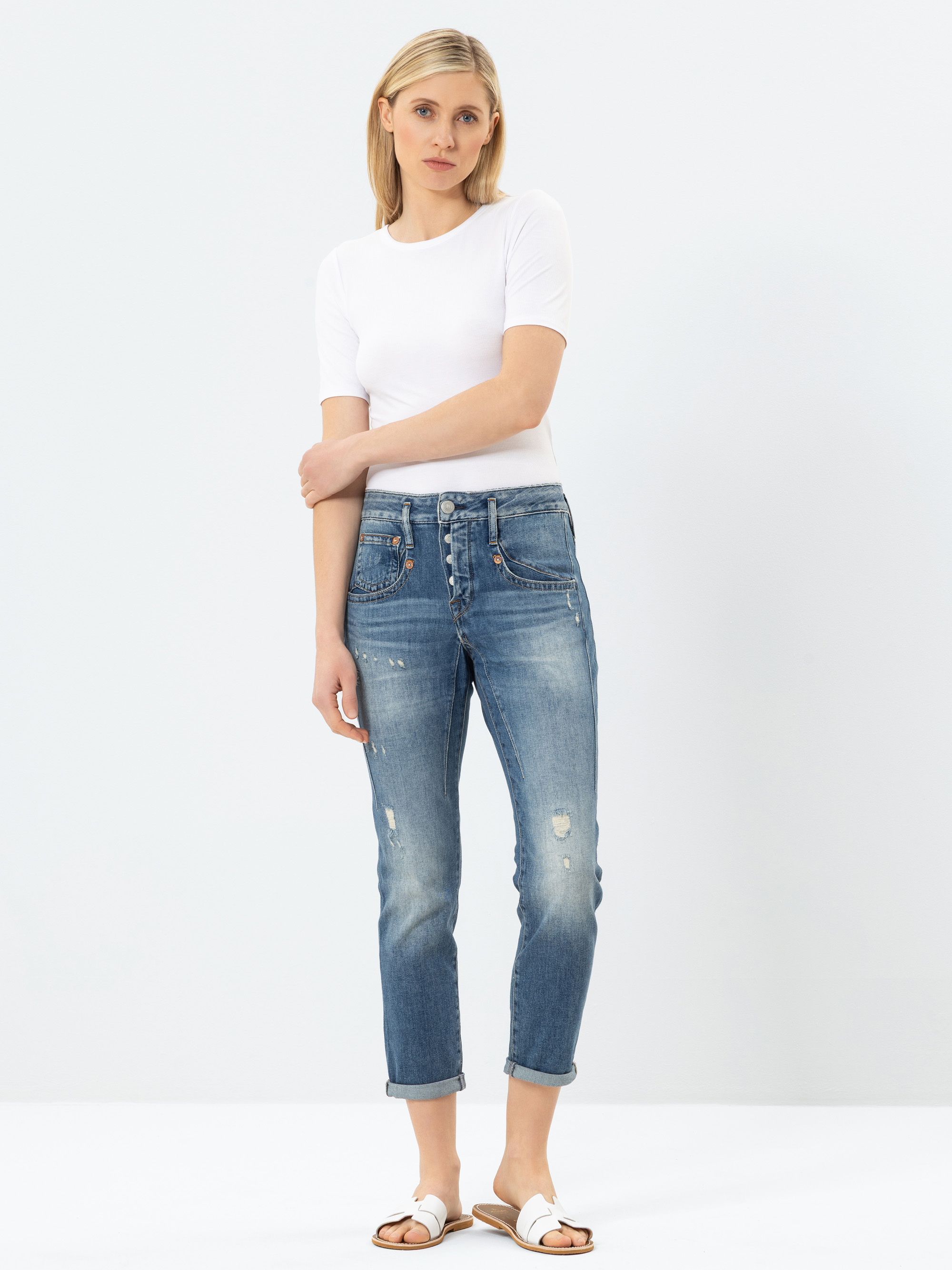 Eine Frau steht vor einem schlichten weißen Hintergrund und trägt ein weißes, kurzärmeliges Oberteil, eine blaue Jeans mit hoher Taille, die mit einem Knopfverschluss versehen ist und leichte Gebrauchsspuren aufweist, sowie weiße Sandalen. Ihre Arme