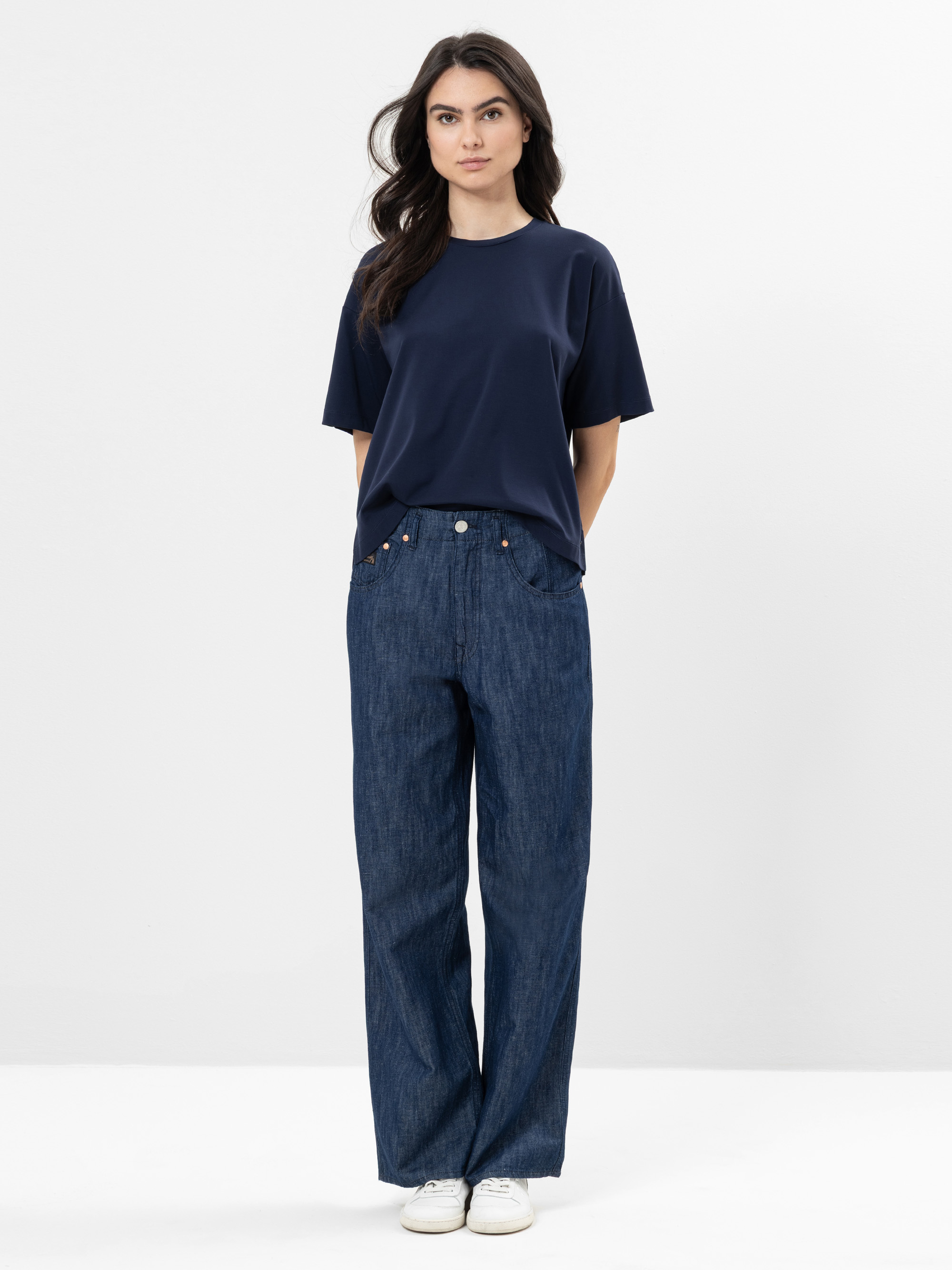 Eine Frau steht vor einem schlichten weißen Hintergrund, trägt ein marineblaues T-Shirt, eine locker sitzende dunkelblaue Jeans und weiße Turnschuhe, hat die Hände auf dem Rücken und einen neutralen Gesichtsausdruck.