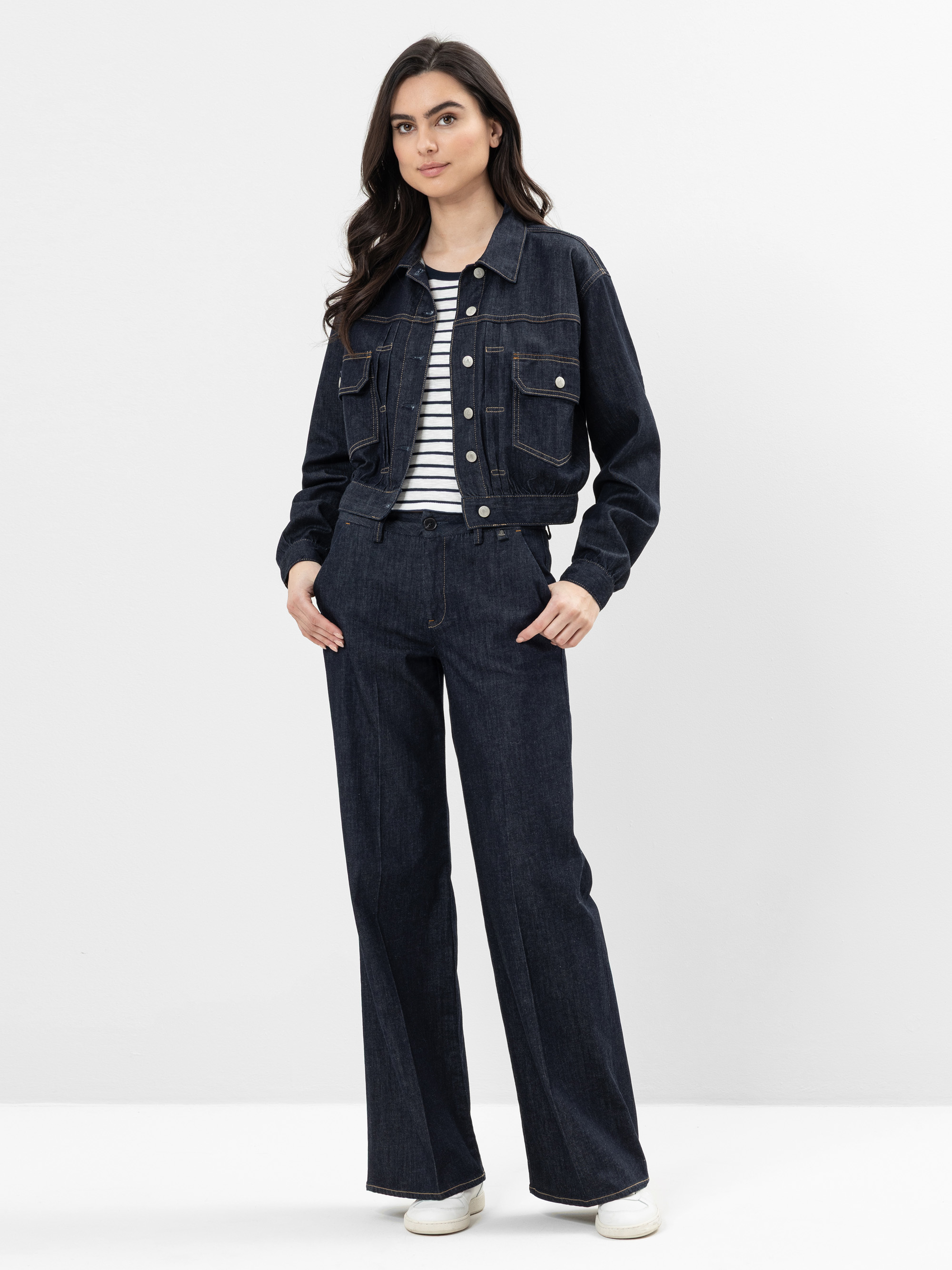 Eine Frau steht vor einem schlichten Hintergrund und trägt eine dunkle Jeansjacke und eine dazu passende Jeans mit weitem Bein über einem schwarz-weiß gestreiften Hemd. Sie hat lange dunkle Haare und trägt weiße Turnschuhe.