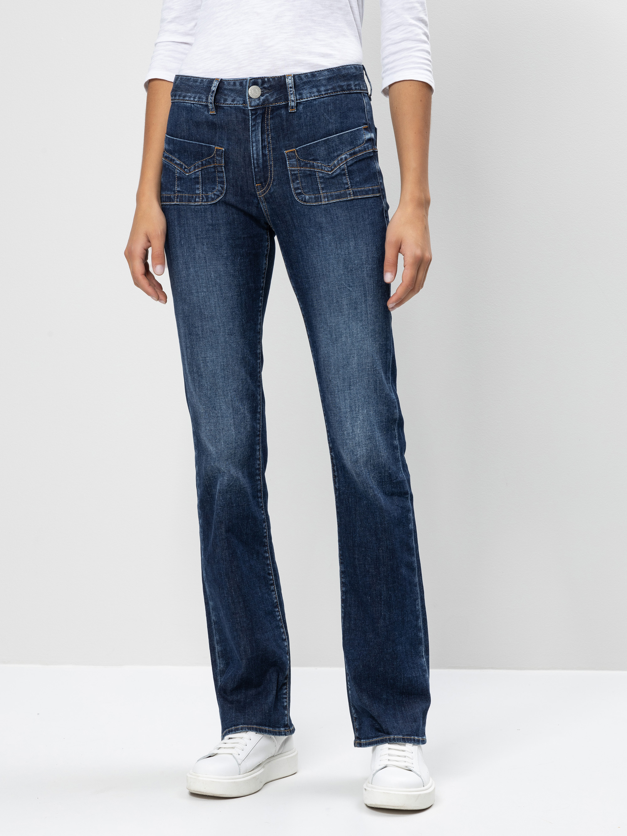 Eine Person, die dunkelblaue Jeans mit geradem Bein und aufgesetzten Vordertaschen, ein weißes langärmeliges Oberteil und weiße Turnschuhe trägt, steht vor einem einfarbigen hellgrauen Hintergrund.