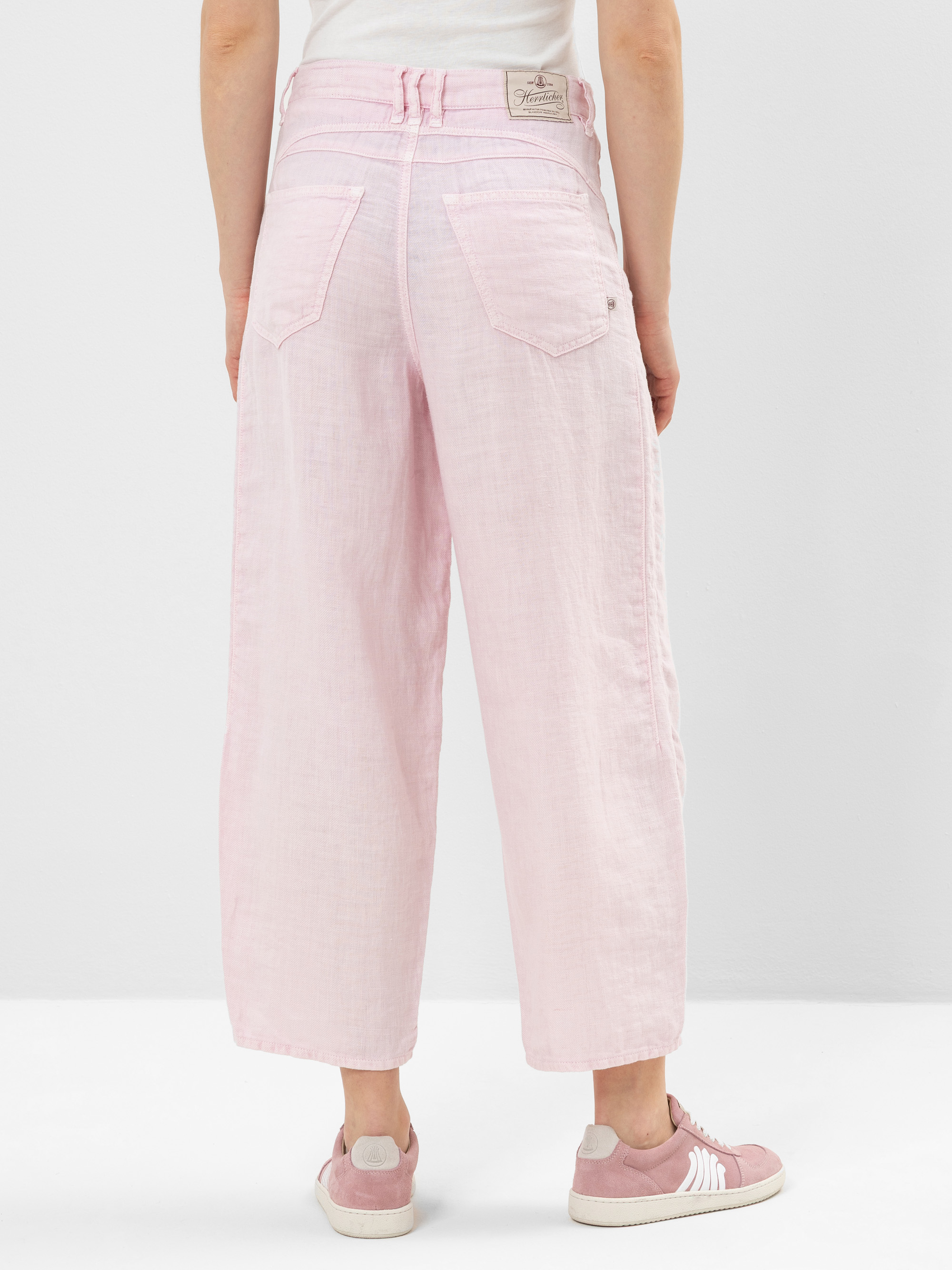 Eine Person steht mit dem Gesicht nach hinten und trägt eine hellrosa Hose mit weitem Bein und dazu passende rosa Turnschuhe. Die Hose hat große Gesäßtaschen und ein weißes Etikett am Bund. Der Boden und der Hintergrund sind weiß.