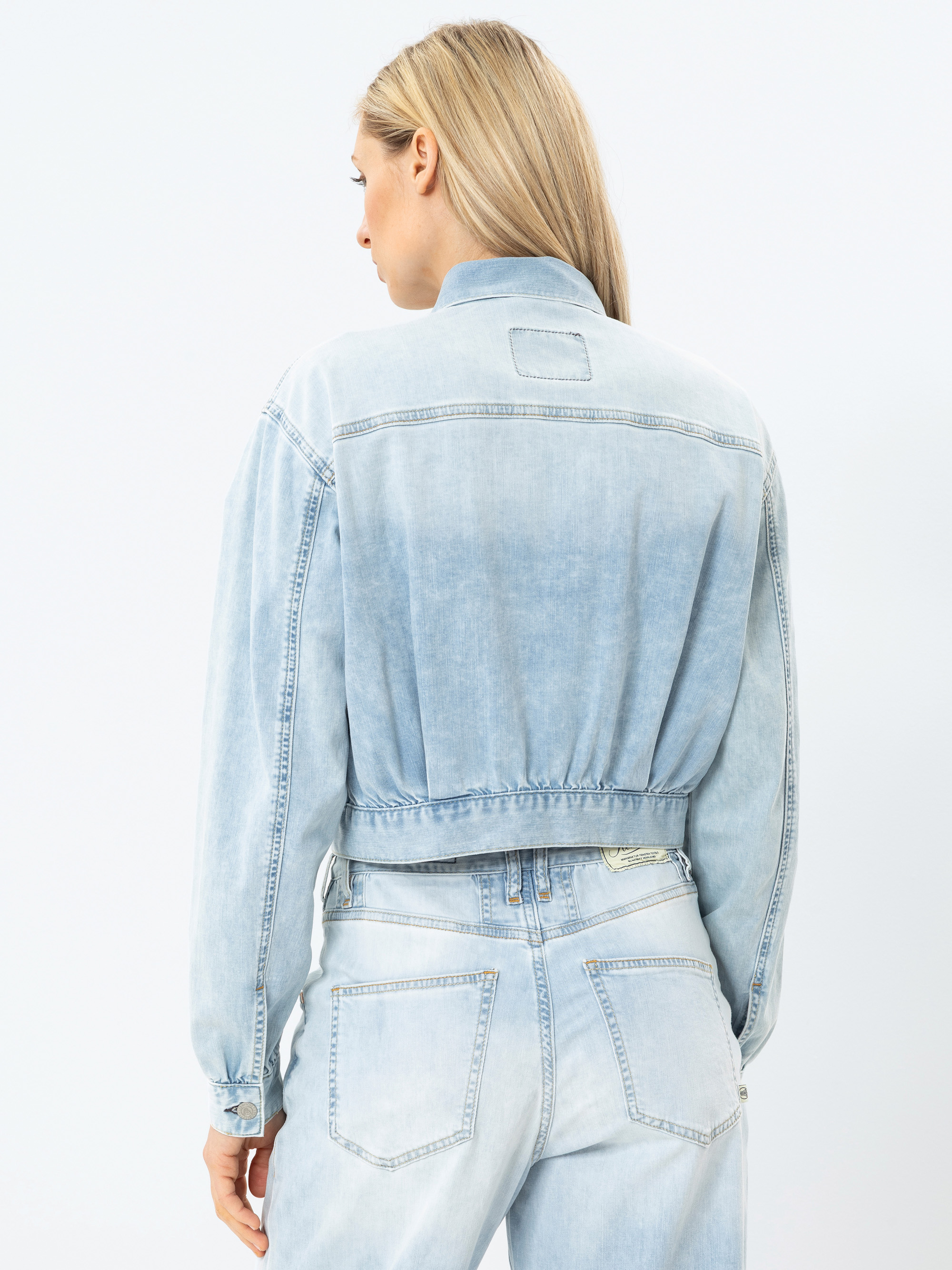 Eine Frau mit langen blonden Haaren ist von hinten zu sehen. Sie trägt eine hellblaue Jeansjacke und dazu passende Jeans mit hoher Taille. Die Jacke hat einen elastischen Bund und sitzt locker. Der Hintergrund ist einfarbig weiß.