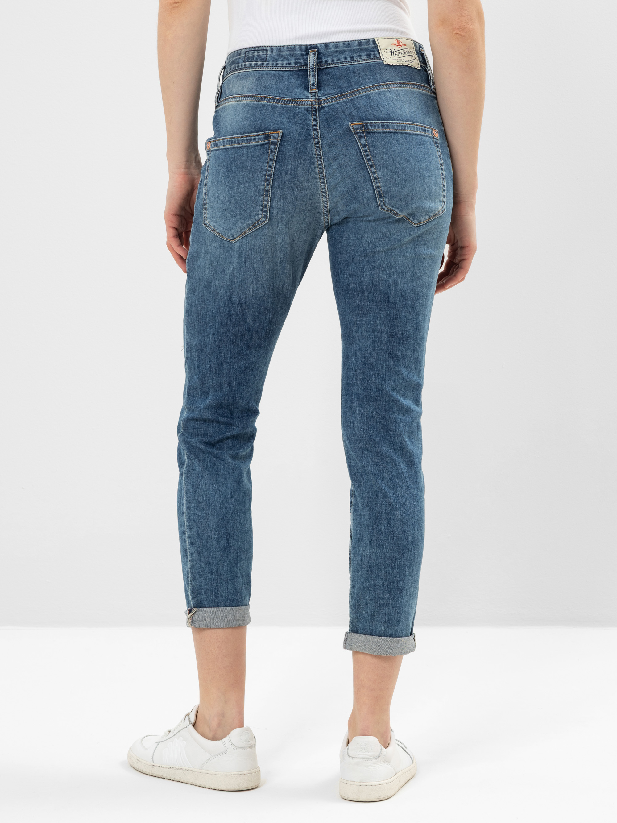 Eine Person, die ein weißes Hemd, eine Jeans mit blauen Bündchen und weiße Turnschuhe trägt, steht mit dem Rücken zur Kamera vor einem schlichten weißen Hintergrund.