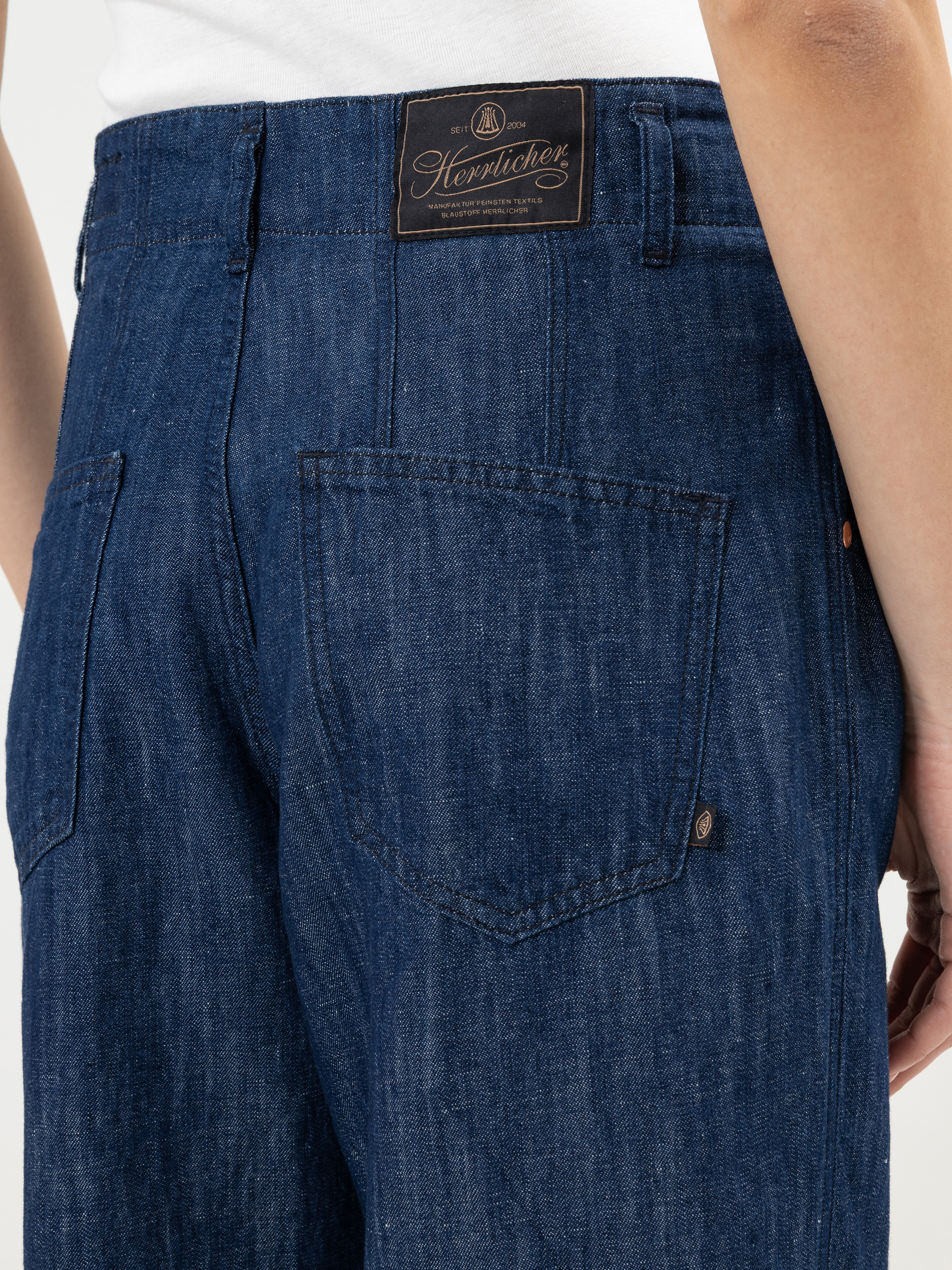 Nahaufnahme einer Person, die eine blaue Jeans trägt. Man sieht die Gesäßtaschen und ein braunes Heimlicher-Markenetikett am Bund. Der rechte Arm der Person ist teilweise sichtbar.