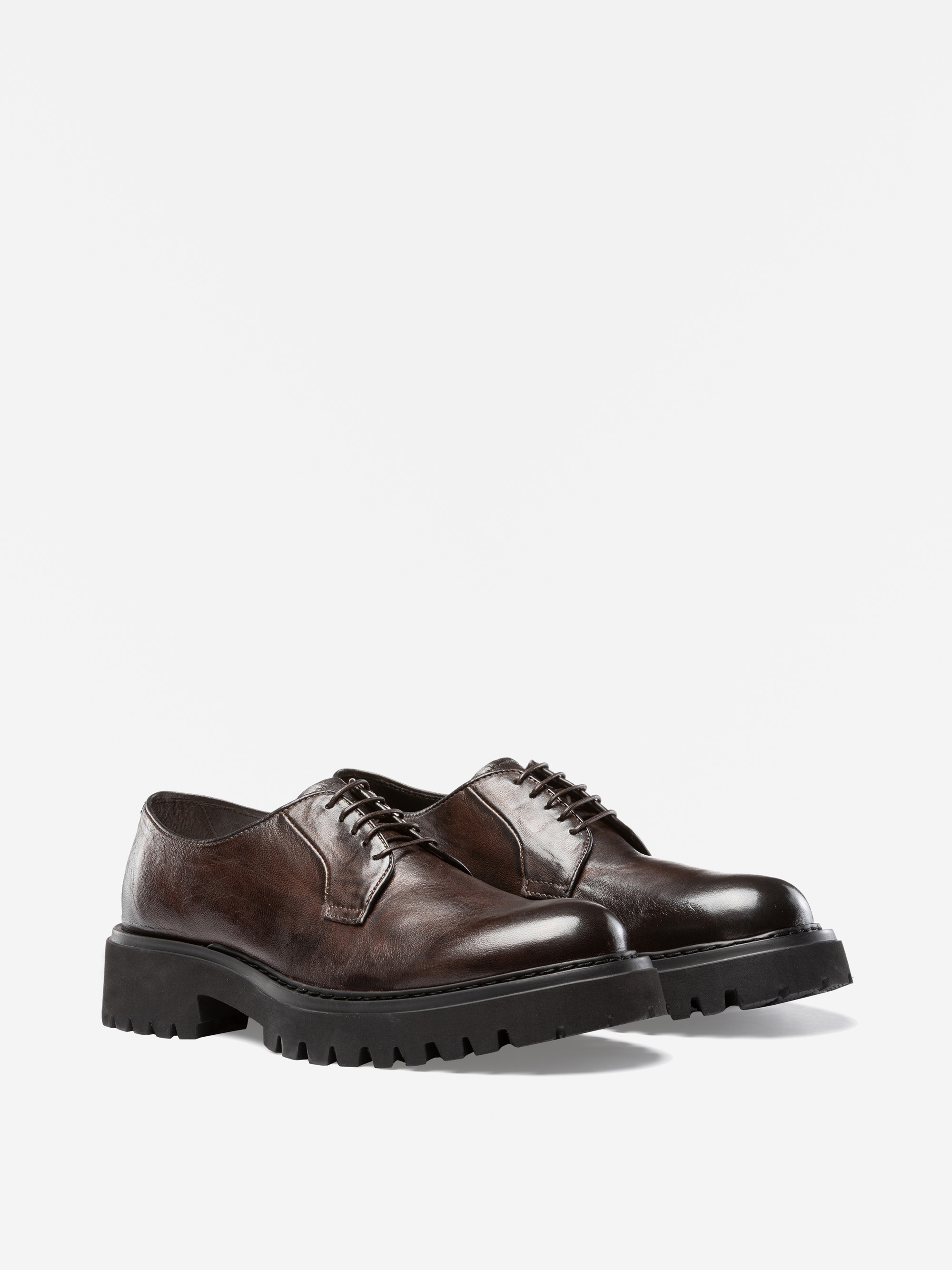 Ein Paar dunkelbraune Lederschnürschuhe mit klobigen schwarzen Gummisohlen auf weißem Hintergrund.