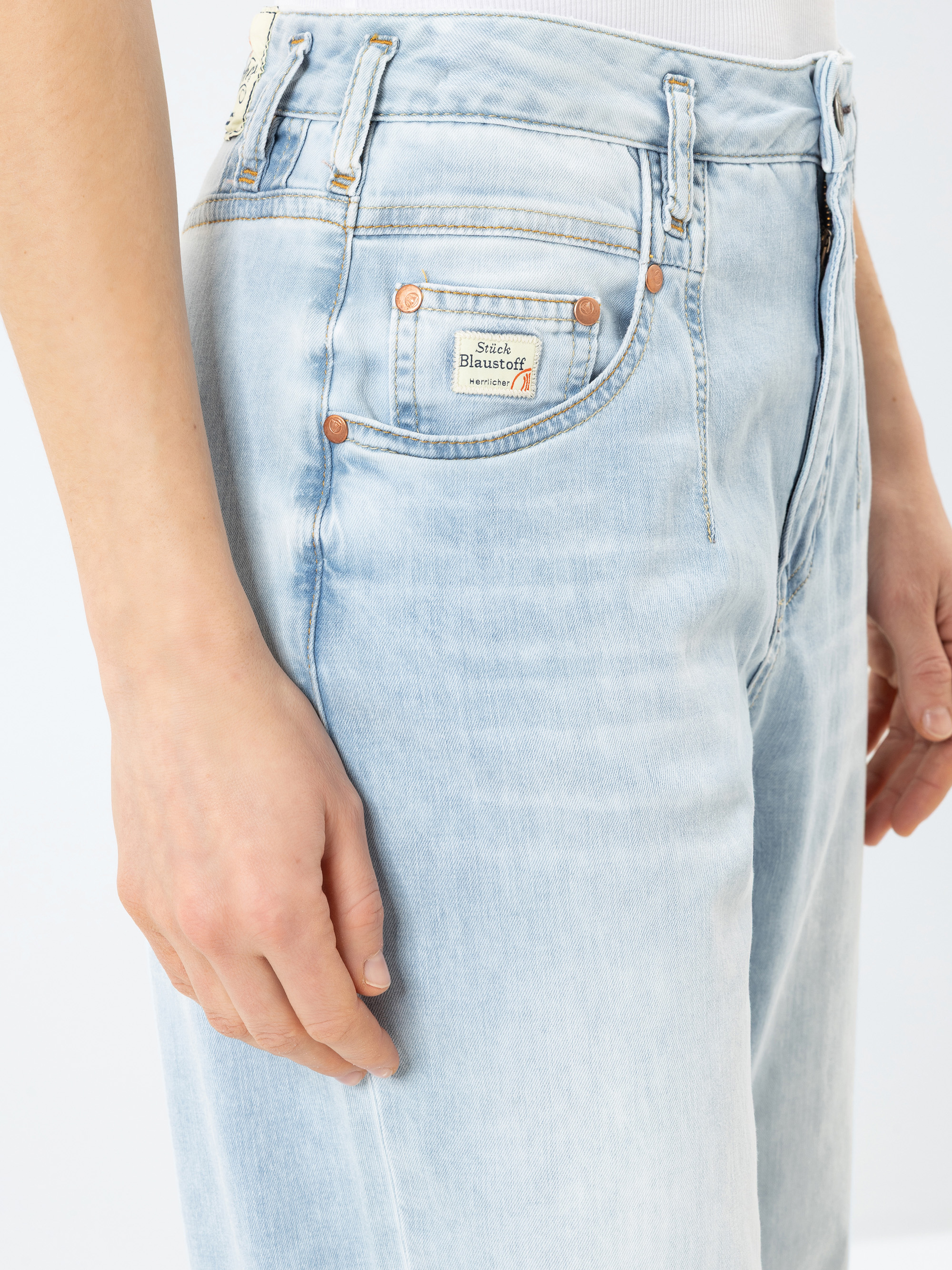 Nahaufnahme einer Person, die eine hellblaue Jeans mit hoher Taille und ein weißes Oberteil trägt, wobei der vordere Taschenbereich und die linke Hand der Person zu sehen sind.