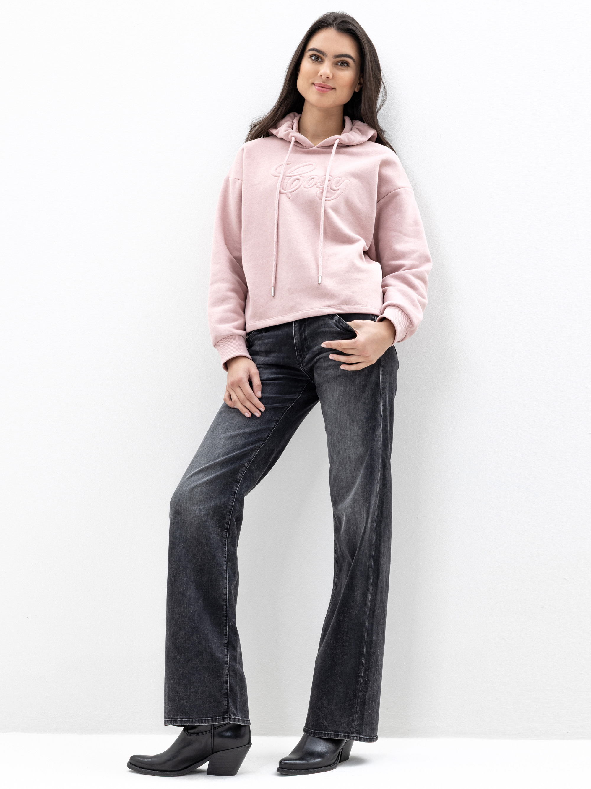Eine Frau steht vor einer weißen Wand und trägt einen hellrosa Kapuzenpulli mit der Aufschrift "Love", dunkle Jeans mit weitem Bein und schwarze Stiefeletten. Sie hat langes dunkles Haar und lächelt sanft, eine Hand in der Tasche.