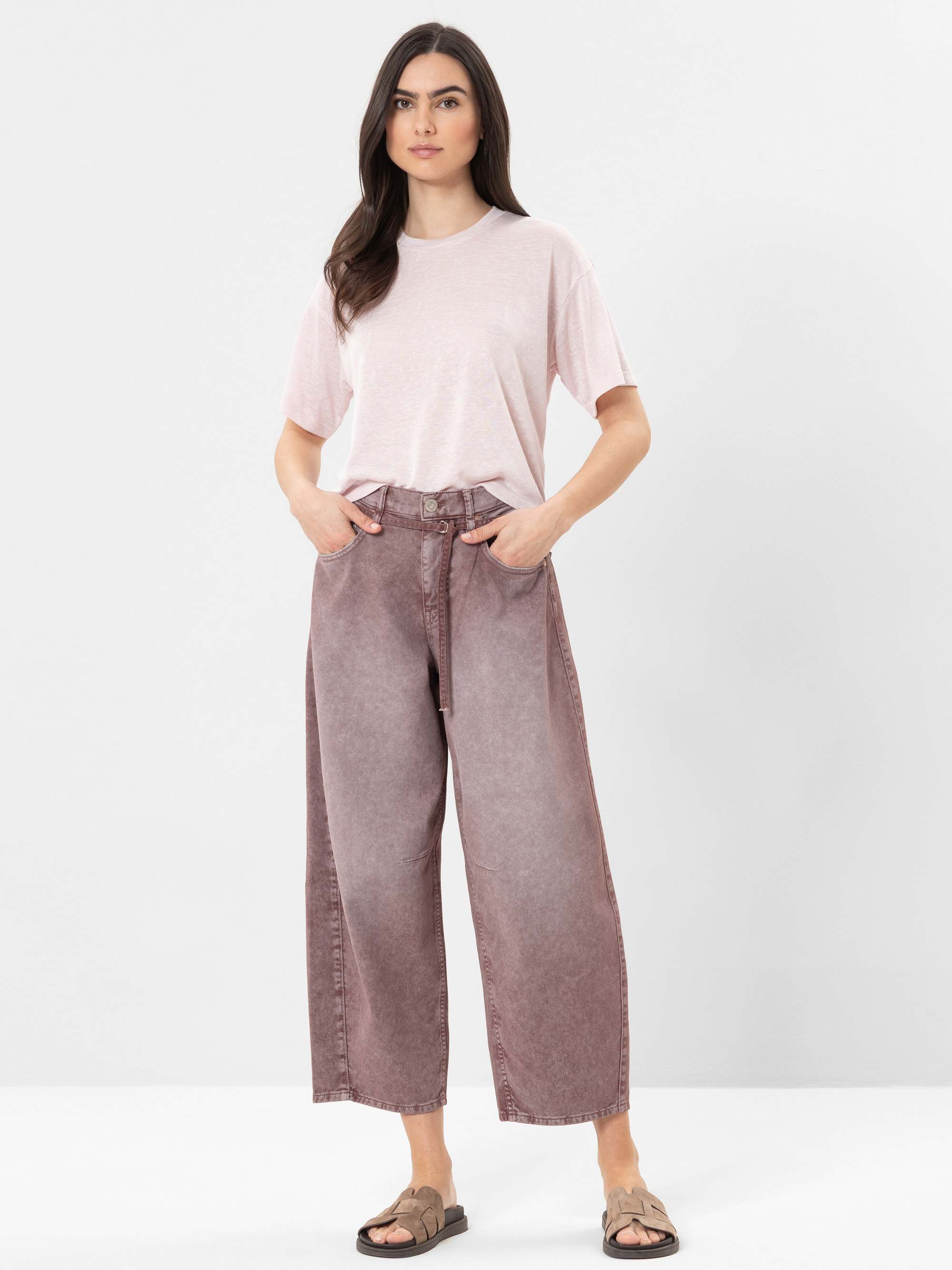 Eine Frau steht vor einem schlichten Hintergrund und trägt ein hellrosa T-Shirt, weite, hochgekrempelte lilafarbene Jeans und beigefarbene Sandalen. Sie hat langes dunkles Haar, die Hände in den Taschen und einen neutralen Gesichtsausdruck.