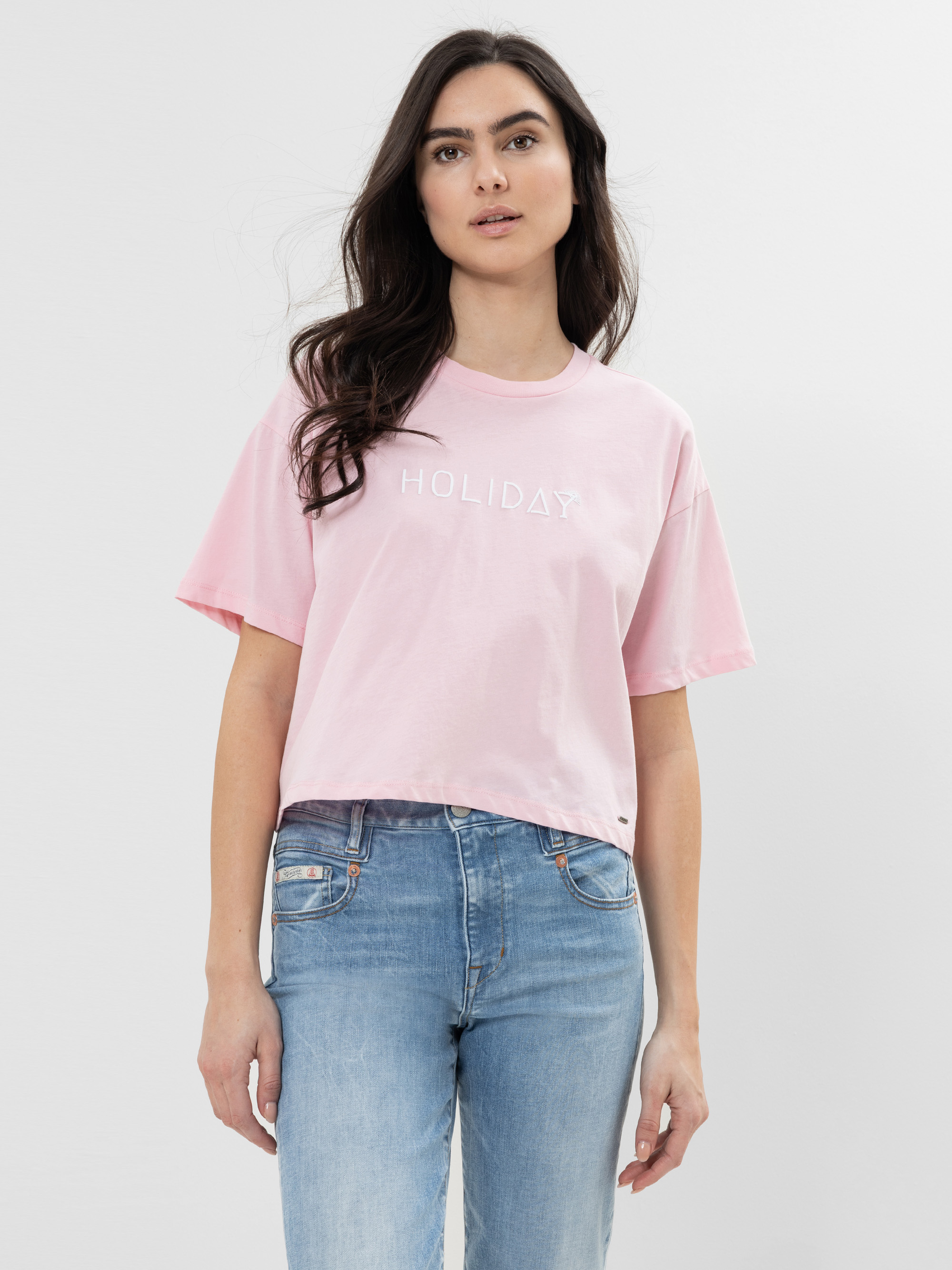 Eine Frau mit langen dunklen Haaren trägt ein hellrosa T-Shirt mit dem Aufdruck HOLIDAY und hellblaue Jeans und steht vor einem schlichten weißen Hintergrund.