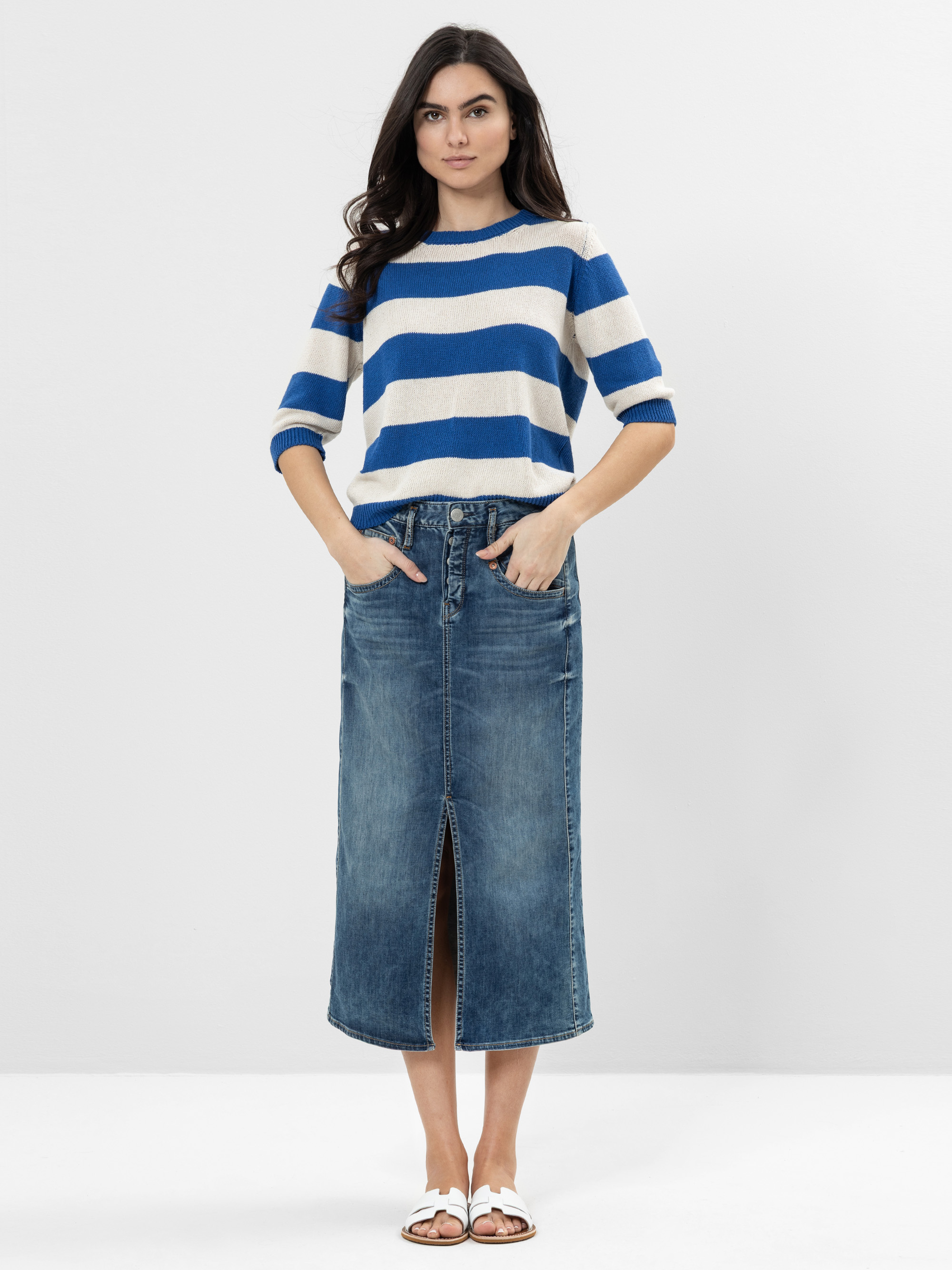 Eine Frau steht vor einem unifarbenen Hintergrund und trägt einen blau-weiß gestreiften Pullover, einen hochgeschnittenen Jeans-Midirock mit einem vorderen Schlitz und weiße Sandalen. Ihre Hände stecken in den Rocktaschen und sie blickt nach vorne.