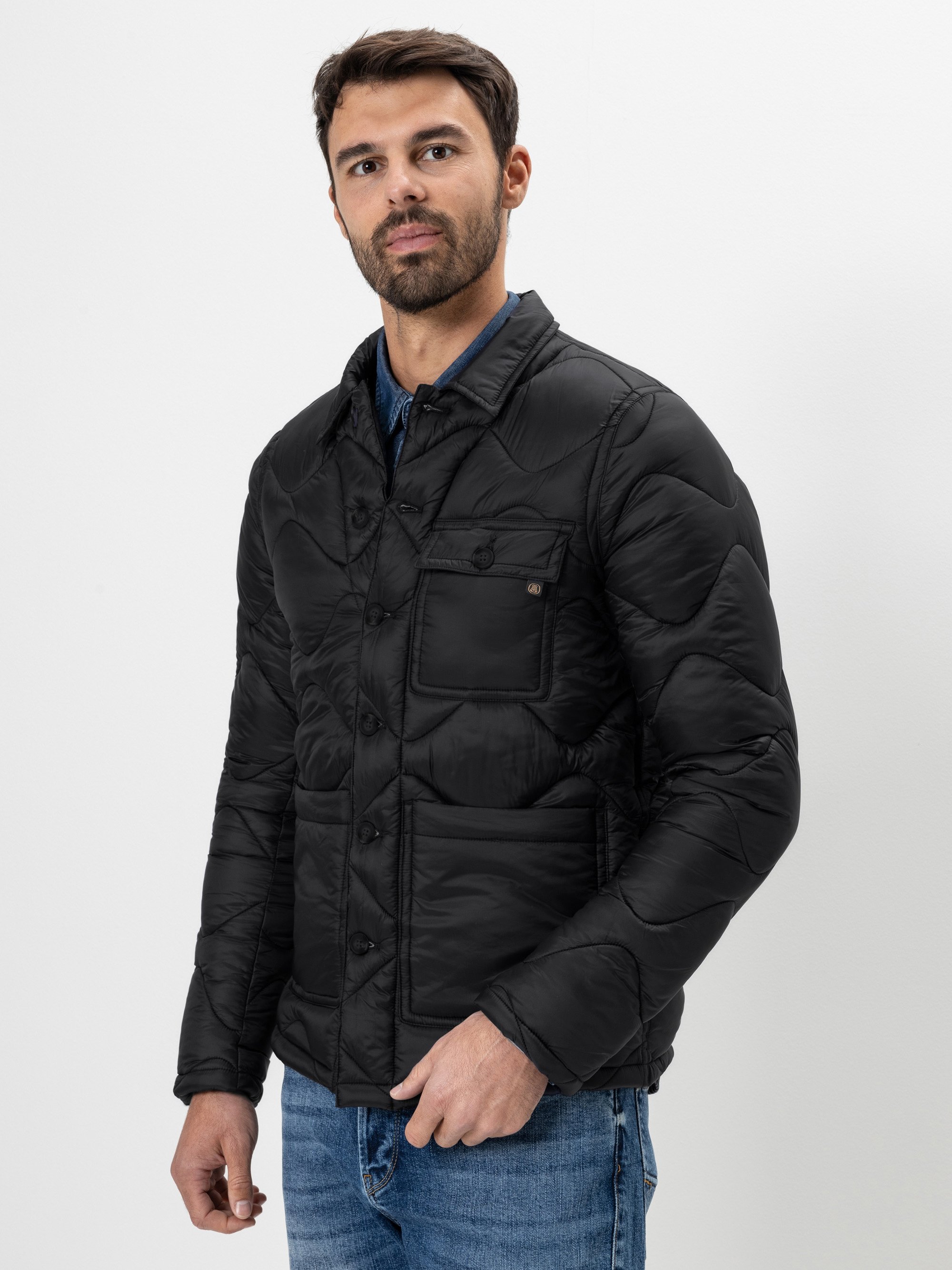 Ein Mann mit kurzen dunklen Haaren und einem Bart trägt eine schwarze Steppjacke über einem blauen Hemd und blauen Jeans. Er steht vor einem schlichten weißen Hintergrund und blickt leicht zur Seite.