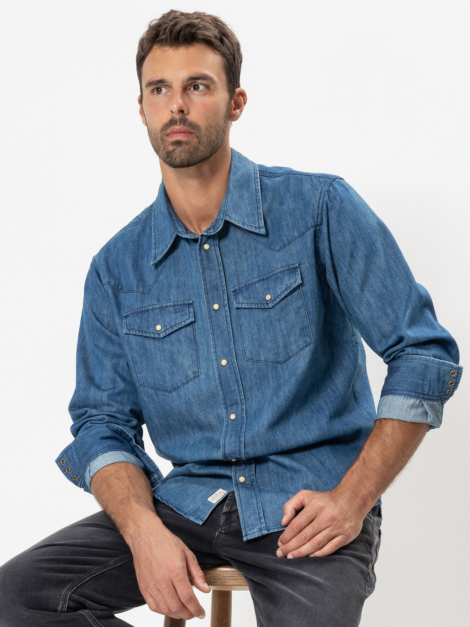 Ein Mann mit kurzen dunklen Haaren und Bart sitzt auf einem Hocker. Er trägt ein blaues Jeanshemd mit geknöpften Brusttaschen und dunkle Jeans. Er hat einen neutralen Gesichtsausdruck und blickt leicht zur Seite.