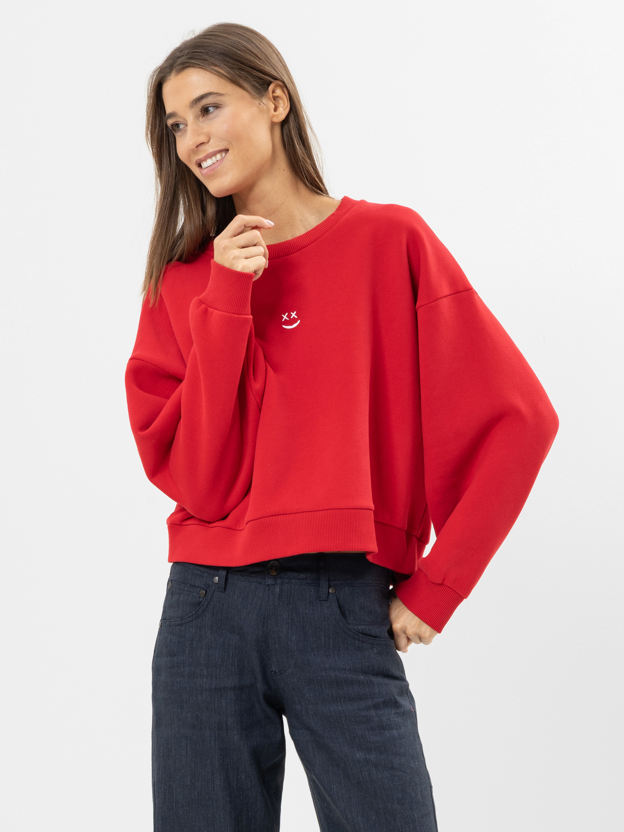 Eine Frau, die ein locker sitzendes rotes Sweatshirt mit einem kleinen weißen Smiley auf der Brust und dunkle Jeans trägt, steht vor einem schlichten weißen Hintergrund, lächelt und schaut zur Seite.