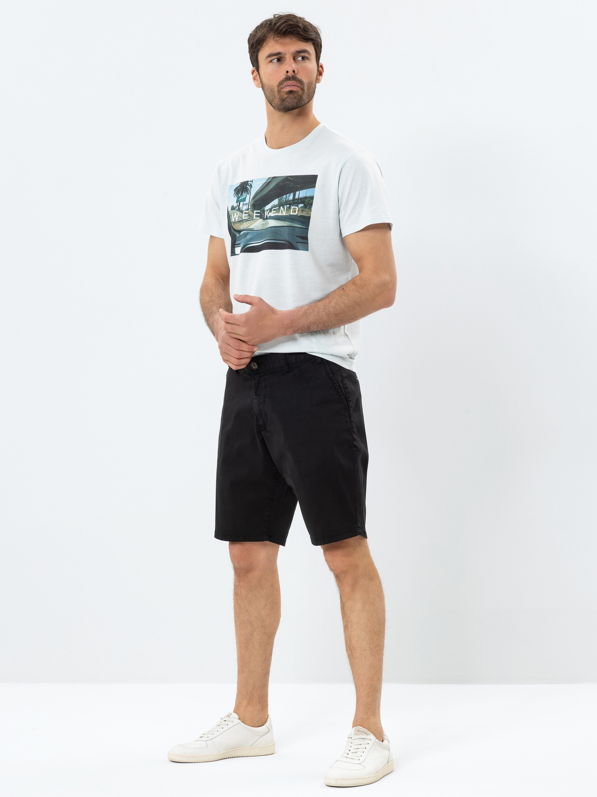 Ein Mann steht vor einem schlichten weißen Hintergrund und trägt ein weißes grafisches T-Shirt, schwarze Shorts und weiße Turnschuhe. Er blickt leicht zur Seite und hat die Hände vor sich verschränkt.