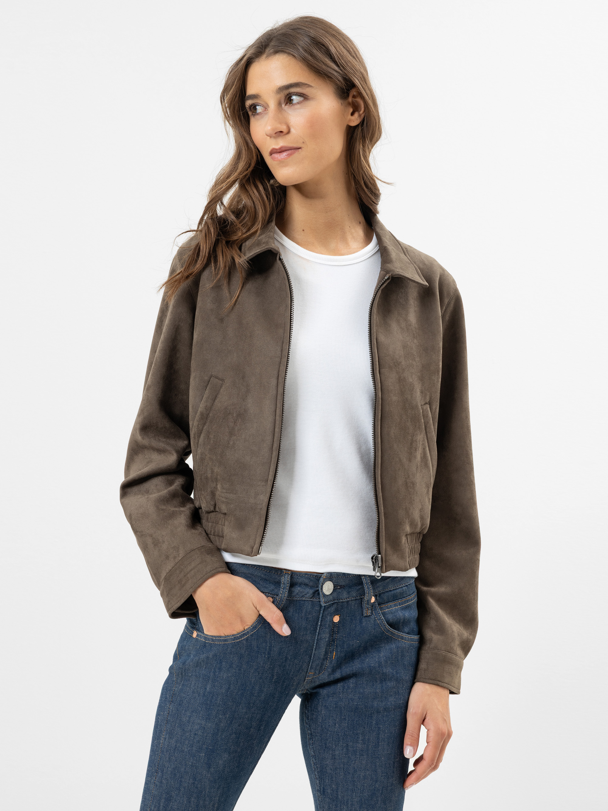 Eine Frau mit langen braunen Haaren trägt eine braune Wildlederjacke über einem weißen Hemd und blaue Jeans. Sie steht vor einem schlichten weißen Hintergrund und schaut leicht nach links.