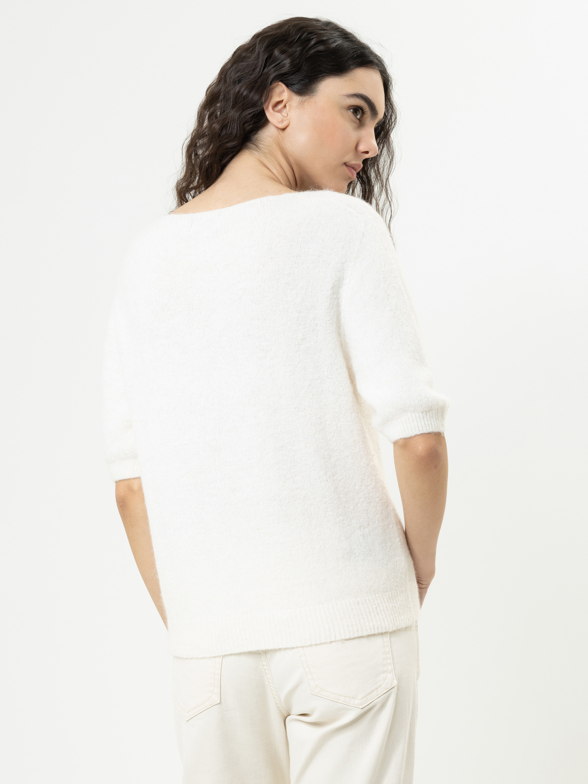 Eine Frau mit langen, gewellten dunklen Haaren steht seitlich und schaut über ihre Schulter. Sie trägt einen weißen, kurzärmeligen Pullover und eine helle Hose vor einem schlichten weißen Hintergrund.