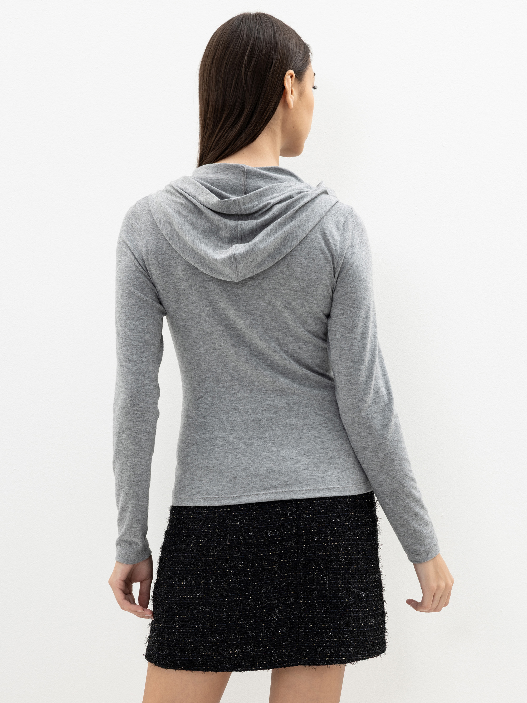 Eine Frau mit langen braunen Haaren, die einen grauen Kapuzenpulli und einen schwarzen, strukturierten Rock trägt, steht mit dem Gesicht zur Kamera vor einem schlichten weißen Hintergrund.