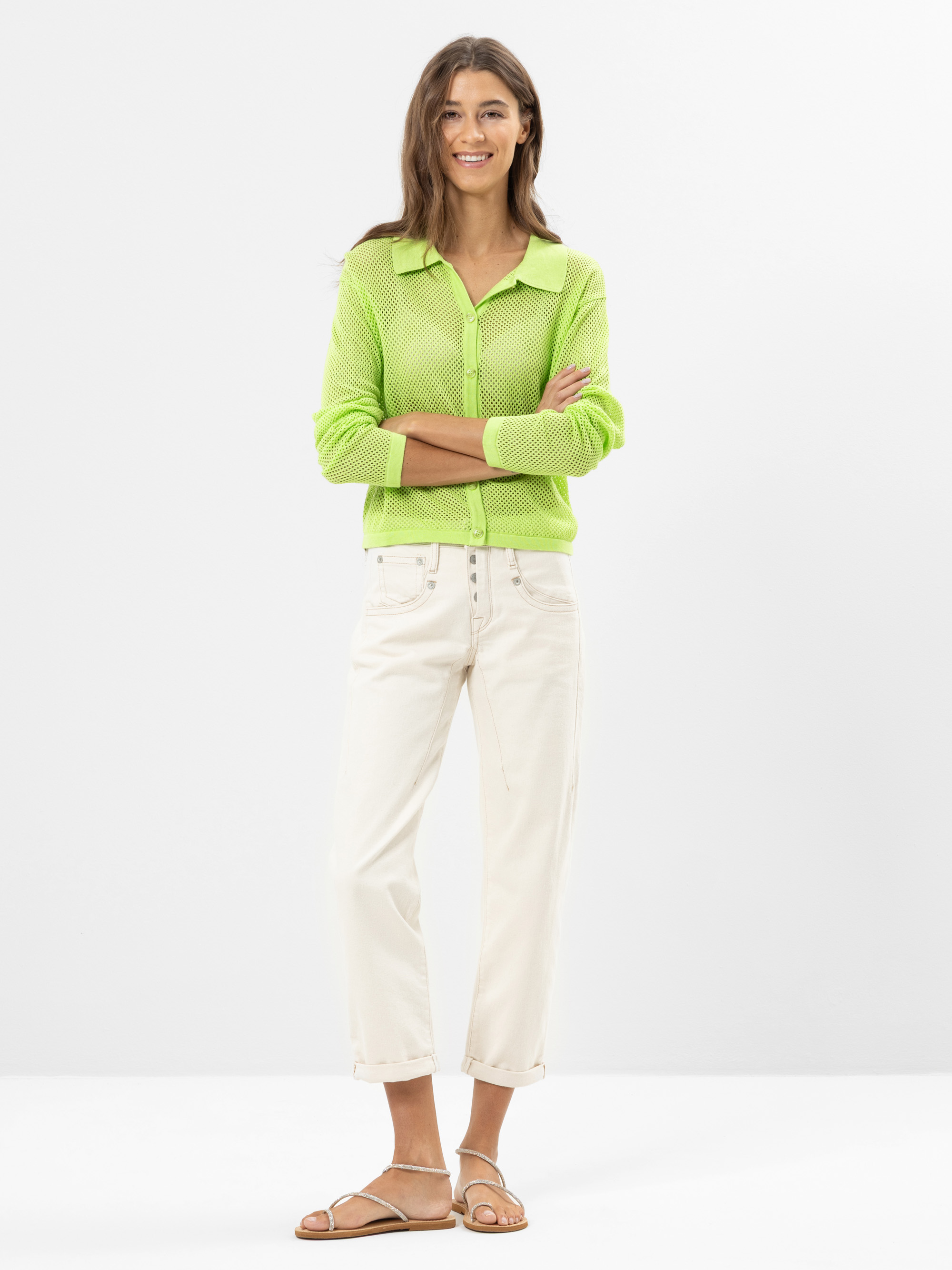 Eine Frau steht lächelnd und mit verschränkten Armen in einem neongrünen Button-up-Hemd, weißen Jeans mit Bündchen und hellbraunen Sandalen vor einem schlichten weißen Hintergrund.