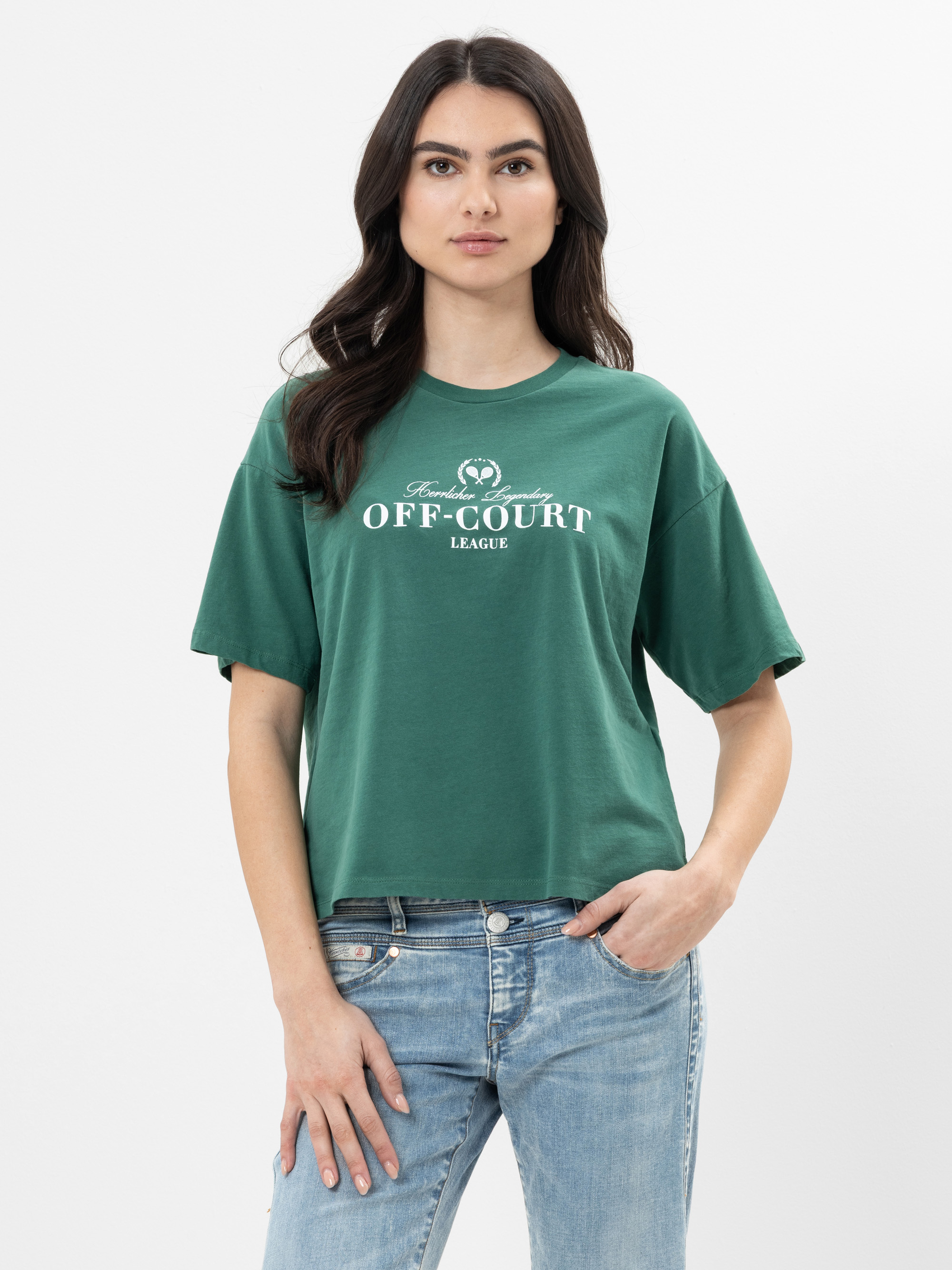 Eine Frau mit langen dunklen Haaren trägt ein grünes OFF-COURT LEAGUE-T-Shirt und blaue Jeans. Sie steht vor einem schlichten weißen Hintergrund und hat eine Hand in der Tasche.