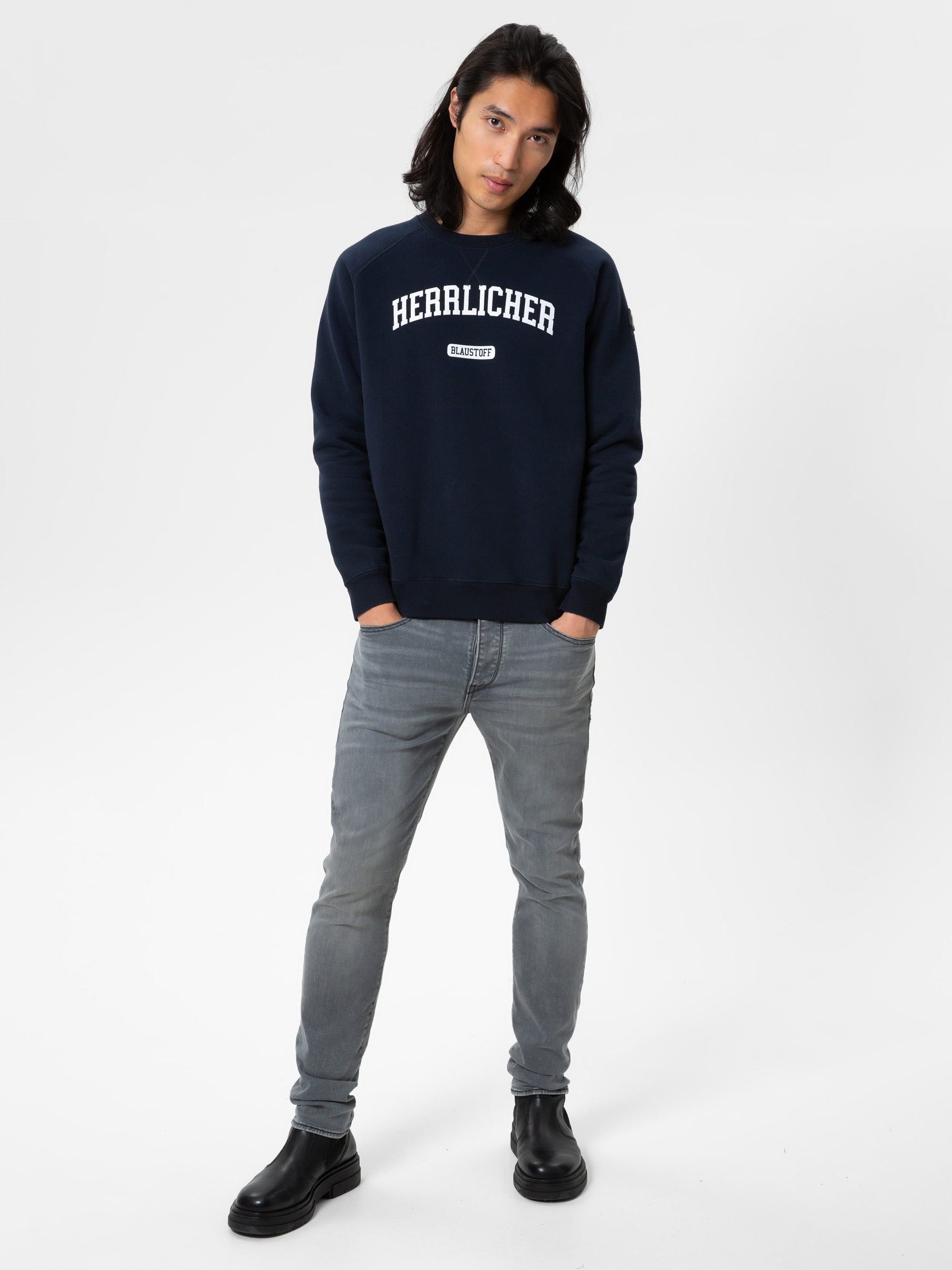 Eine Person mit langen dunklen Haaren steht vor einem schlichten weißen Hintergrund, trägt ein marineblaues Sweatshirt mit der Aufschrift "HERZLICHER", hellgraue Jeans und schwarze Stiefel. Ihre Hände stecken in den Taschen, und sie schaut in die Kamer