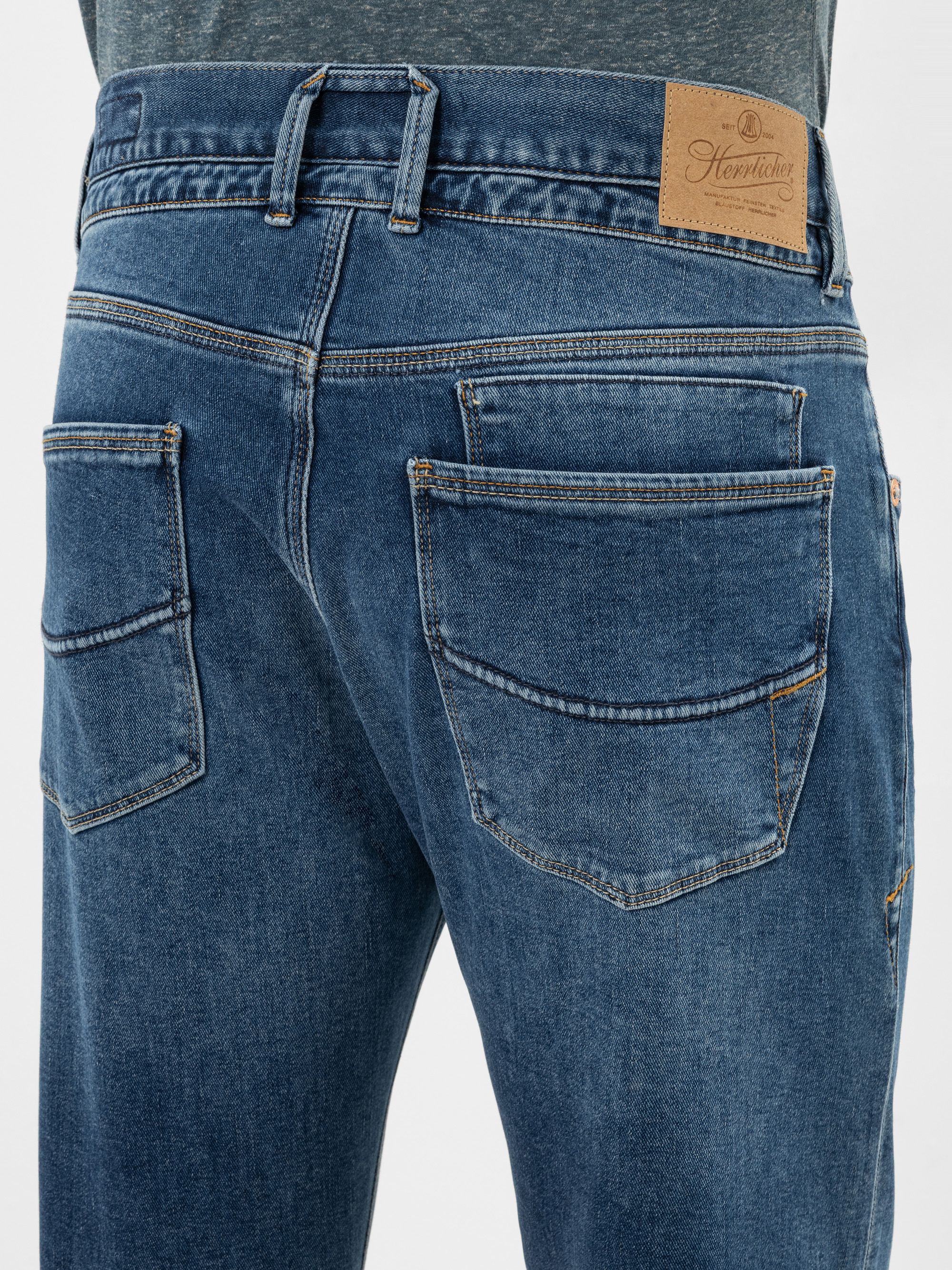 Nahaufnahme des Rückens einer Person, die eine blaue Jeans und ein graues Hemd trägt, mit zwei großen Gesäßtaschen und einem braunen Lederaufnäher am Hosenbund, auf dem die Marke Hawthorn zu sehen ist.