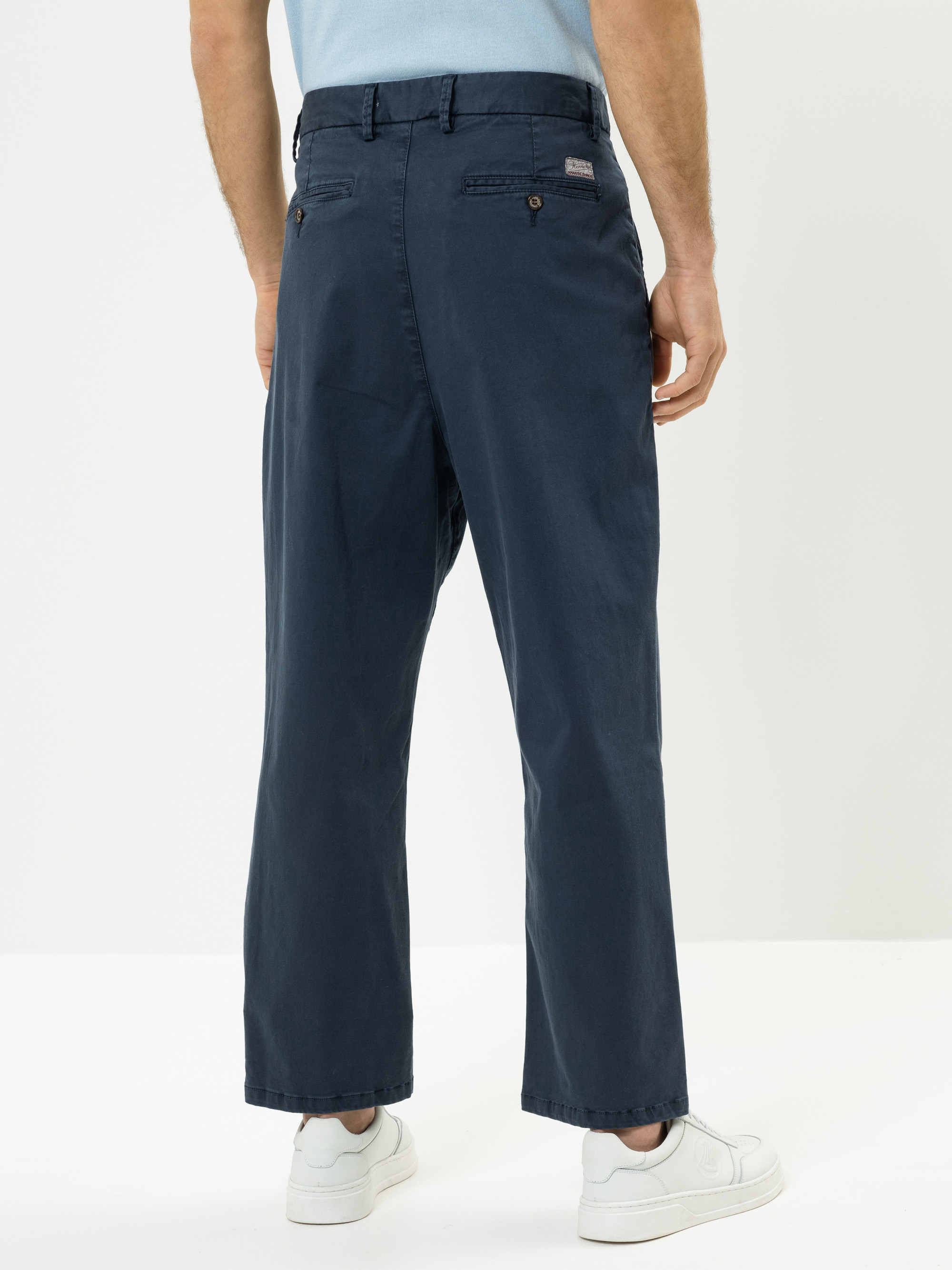Ein Mann steht mit dem Gesicht zur Wand und trägt eine marineblaue Hose mit weitem Bein, ein hellblaues Hemd mit Stecktuch und weiße Turnschuhe. Der Hintergrund ist schlicht und weiß.
