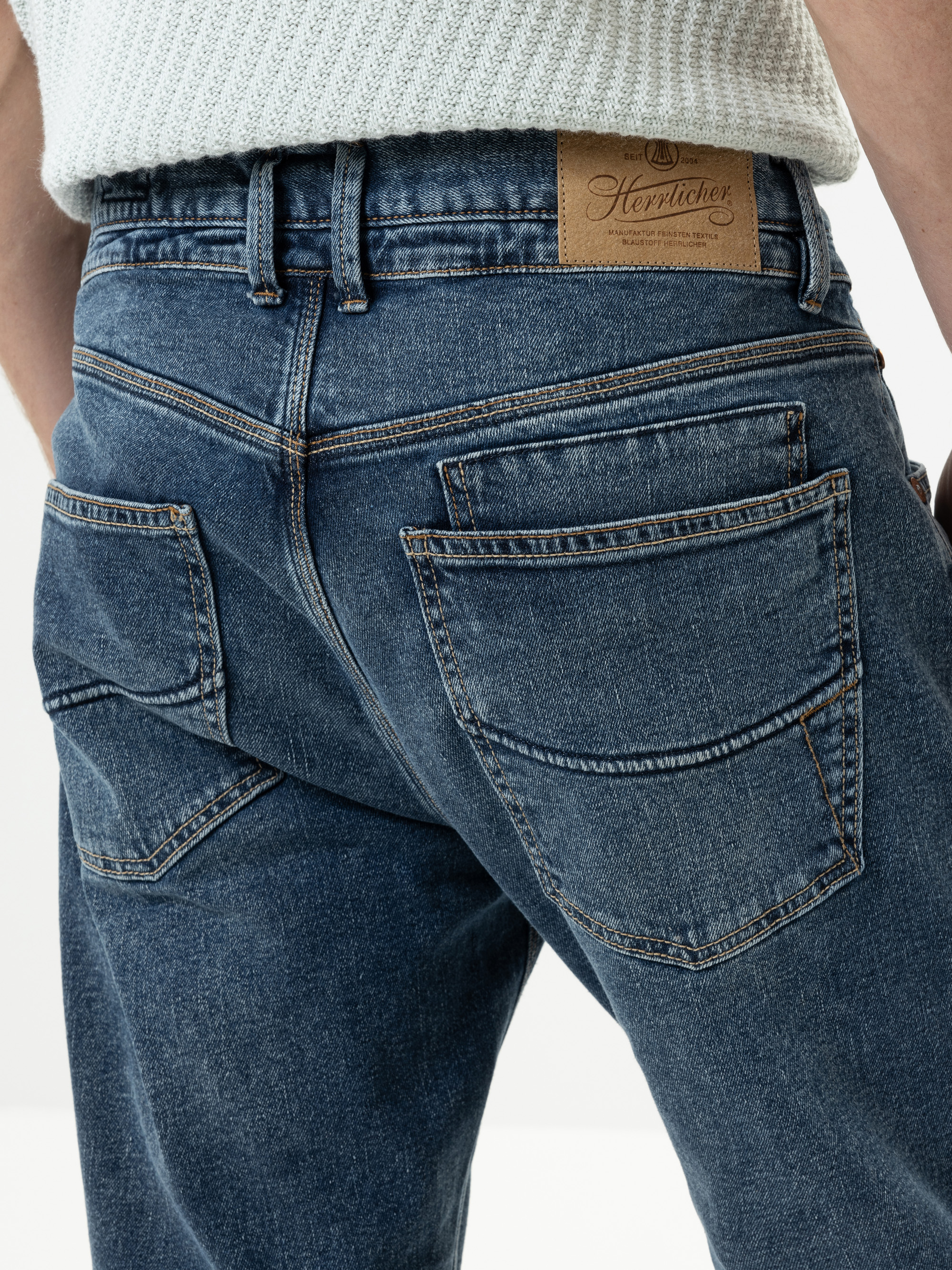 Nahaufnahme einer Person, die eine blaue Jeans und einen hellen Strickpullover trägt, wobei die Gesäßtaschen und ein brauner Markenaufnäher aus Leder am Bund zu sehen sind.