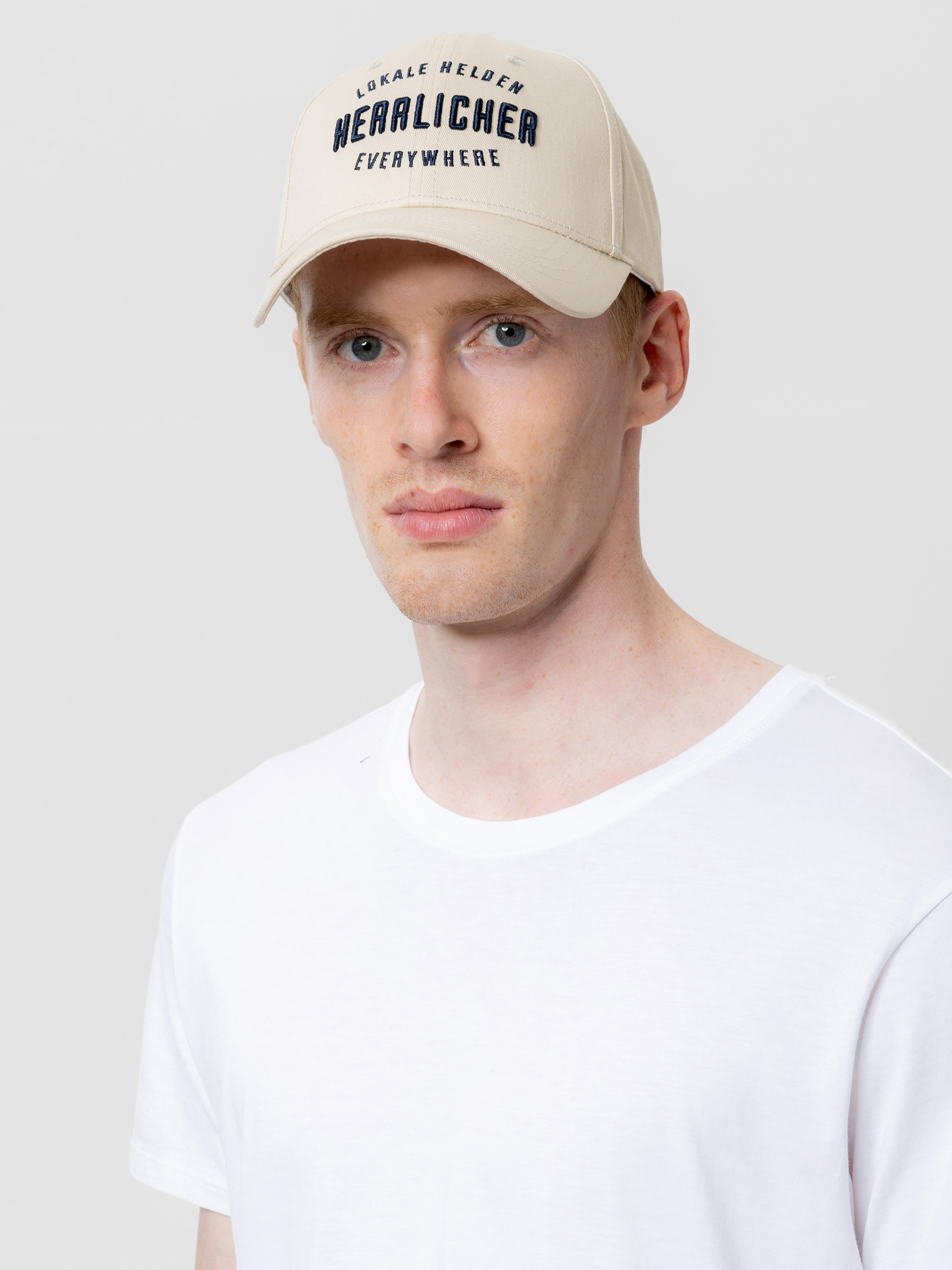 Ein Mann mit heller Haut und kurzen roten Haaren trägt eine beigefarbene Baseballkappe mit der Aufschrift Lokale Helden Herrlicher Everywhere und ein schlichtes weißes T-Shirt vor einem hellgrauen Hintergrund.