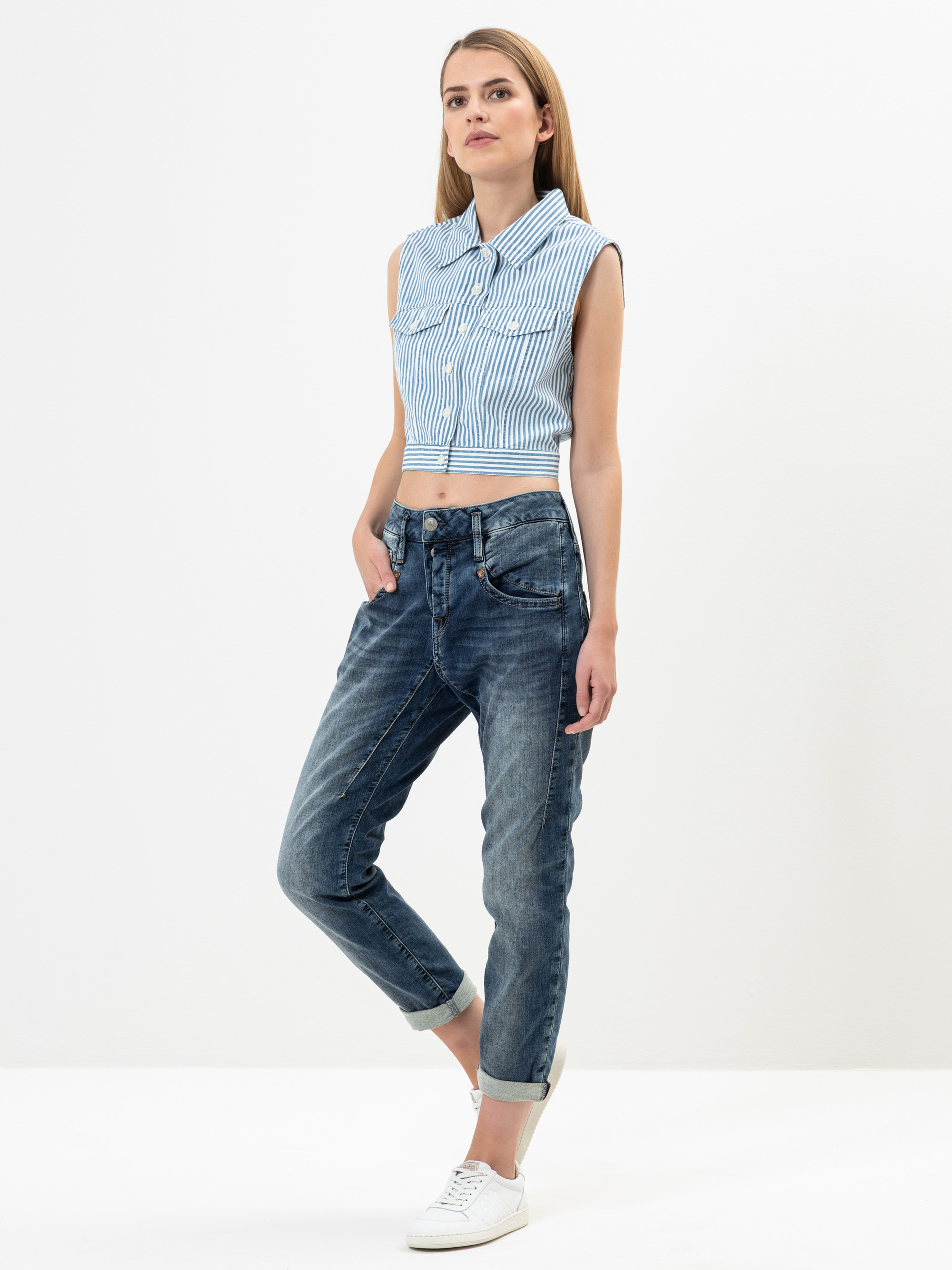 Eine Frau steht vor einem schlichten Hintergrund und trägt ein ärmelloses, blau-weiß gestreiftes, geknöpftes Oberteil, blaue Jeans mit Bündchen und weiße Turnschuhe. Sie hat glattes, hellbraunes Haar und eine entspannte Pose mit einer Hand in der Ta