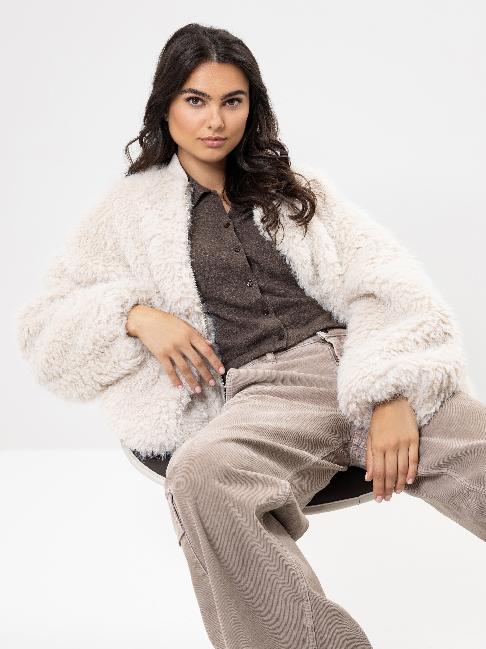 Eine Frau mit langen dunklen Haaren sitzt selbstbewusst auf einem Stuhl und trägt eine flauschige weiße Kunstpelzjacke, ein braunes Button-up-Hemd und eine lockere beigefarbene Kordhose vor einem schlichten weißen Hintergrund.