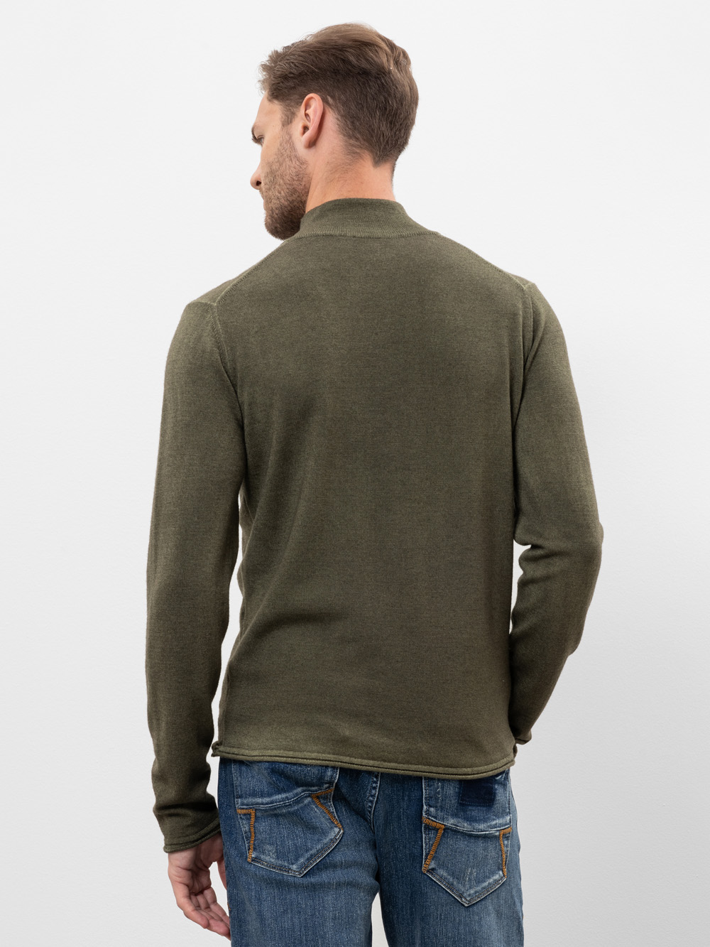 Ein Mann mit kurzen Haaren ist von hinten zu sehen. Er trägt ein langärmeliges olivgrünes Hemd und eine blaue Jeans mit sichtbaren Nähten und steht vor einem schlichten weißen Hintergrund.